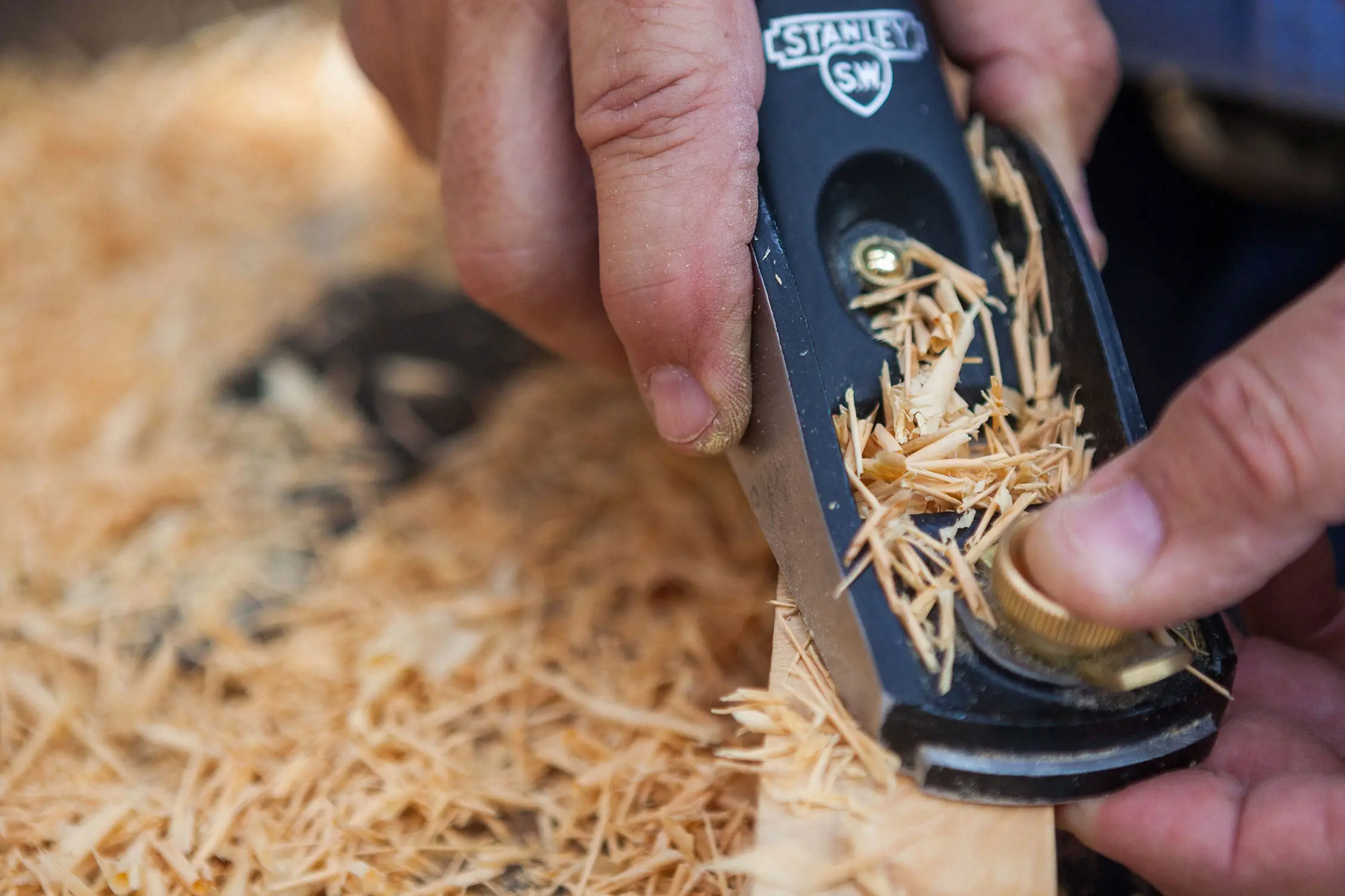 A close-up of a person pushing a wood plane across a piece of timber. Thin splinters of wood shavings cover the plane and the workbench beneath.