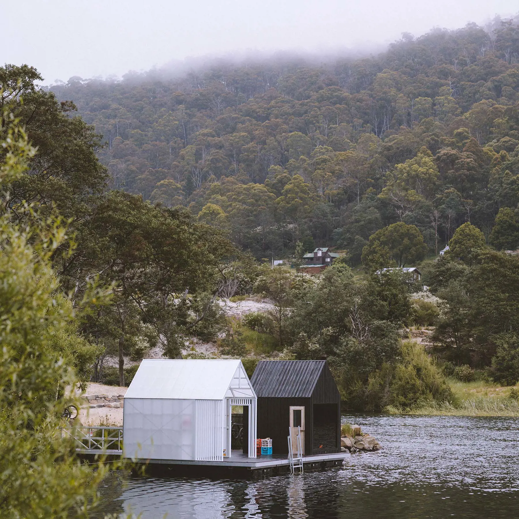 On the edge of a tranquil lake surrounded by misty forested mountains, two small huts sit on a pontoon over the water.