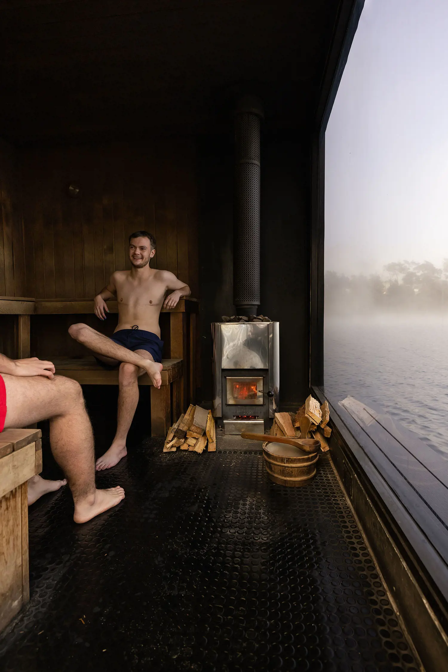 Inside a sauna, two people sit on wooden benches. The sauna stove is lit with stacks of wood next to it. Through the glass wall, a misty lake is visible.