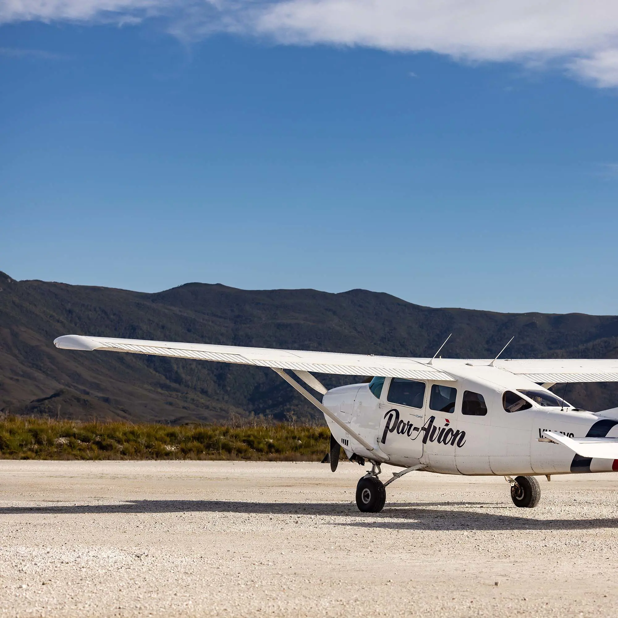 A light plane with 'Par Avion' painted along its body rests on a wide, flat sandy beach. In the background, coastal scrub gives way to steep mountains rising into the clear blue sky.