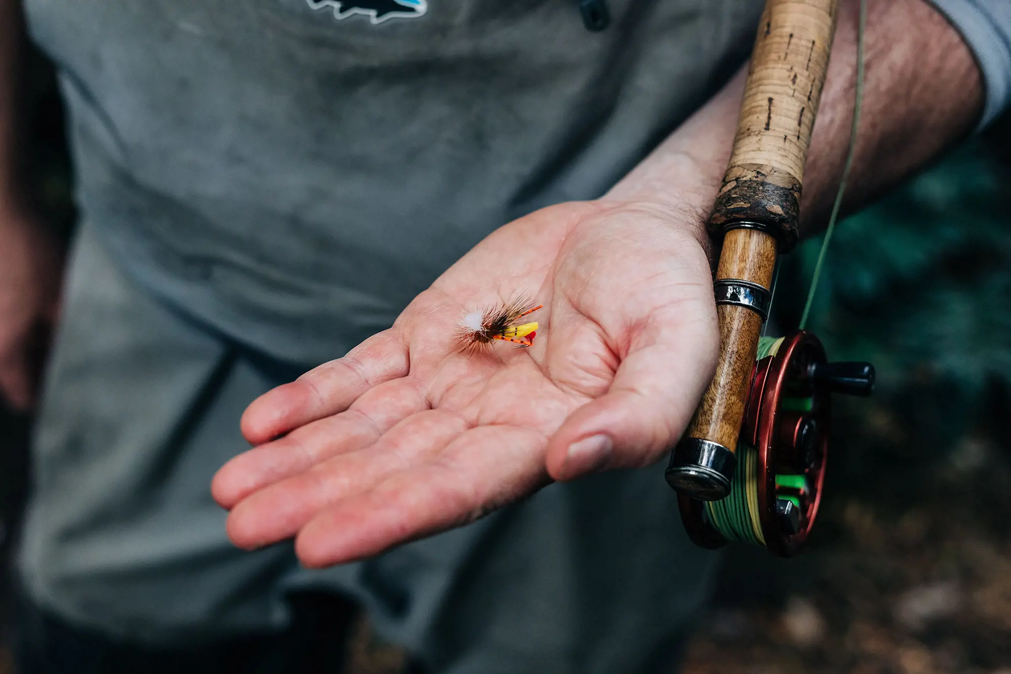 A person holds a small fishing fly in his outstretched hand, and a fly fishing rod is tucked under his arm.