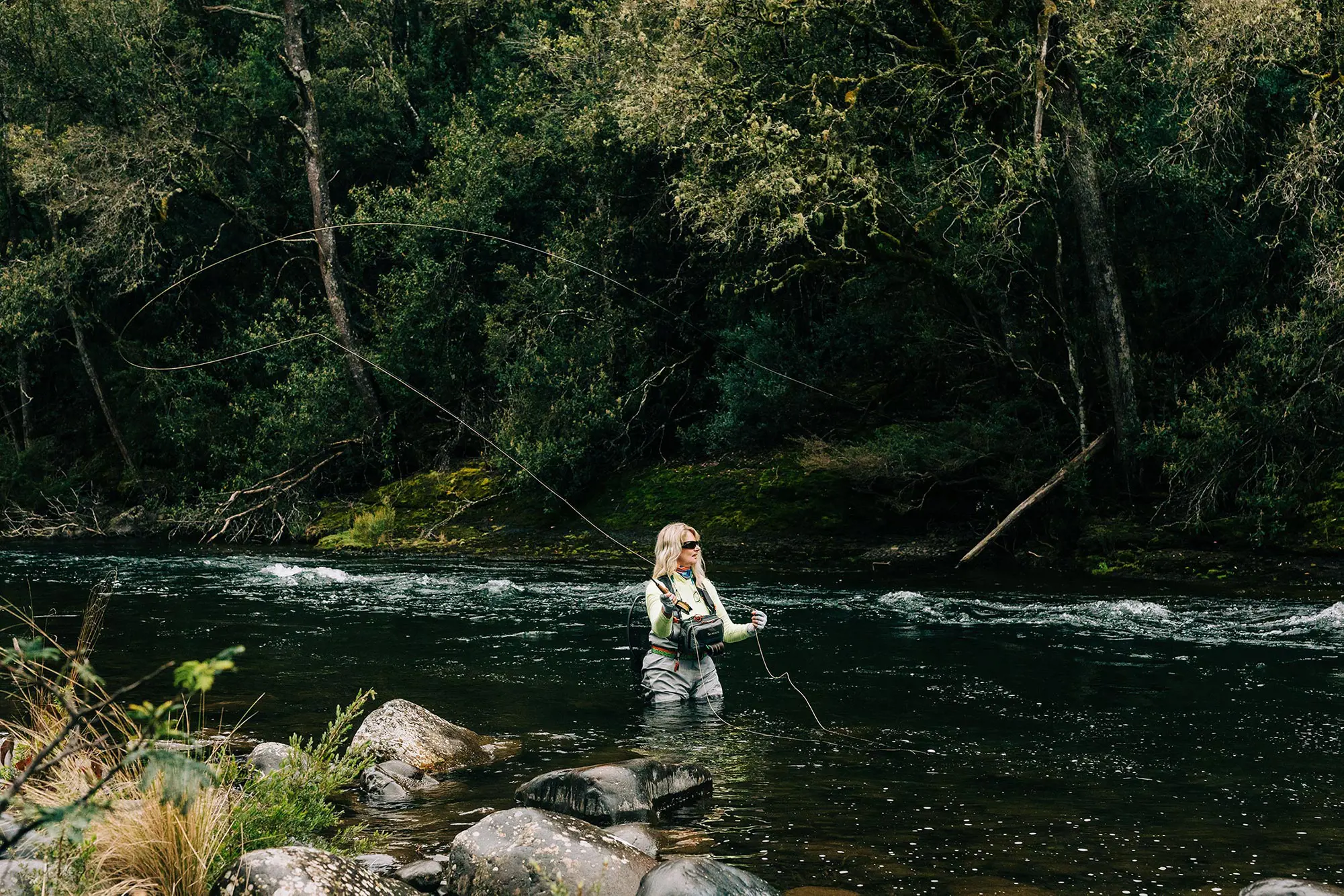 A woman in waders stands in thigh-deep water of a river surrounded by bush, tossing a line. She is wearing dark sunglasses.
