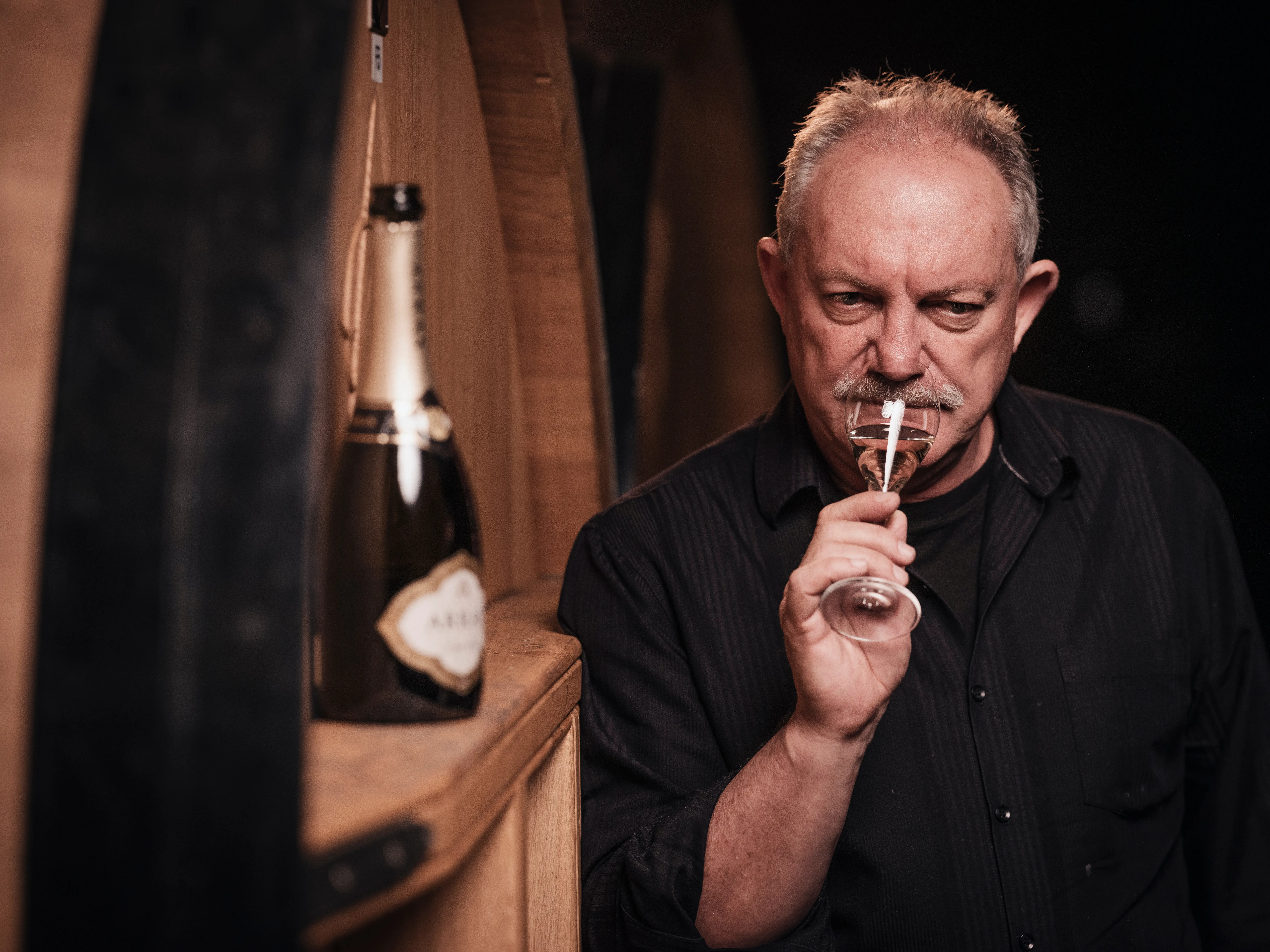 A man in a black shirt holds a glass of champagne up to his nose, standing next to a wooden shelf holding an open champagne bottle.