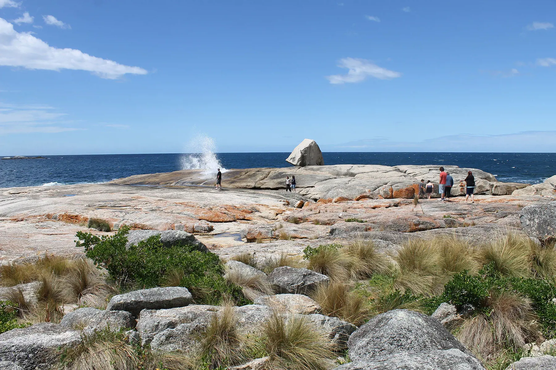 A group of people stand and sit on a rocky shoreline with spraying water shooting up from amid the rocks. The ocean is in the background under a clear blue sky with scattered clouds. 