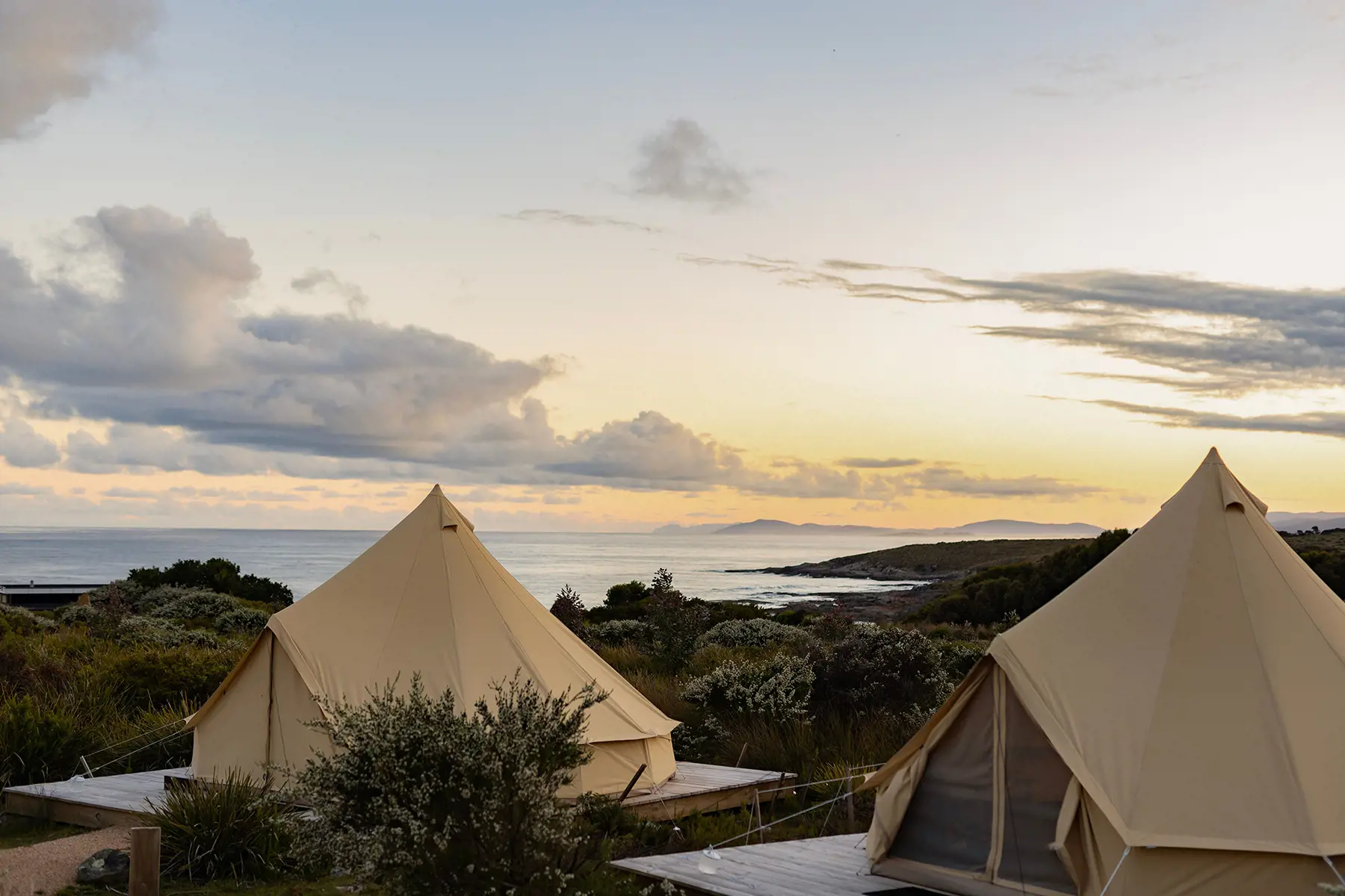 Two beige canvas tents are set up on wooden platforms in a coastal landscape at sunset. The ocean and distant hills are visible under a partly cloudy sky.