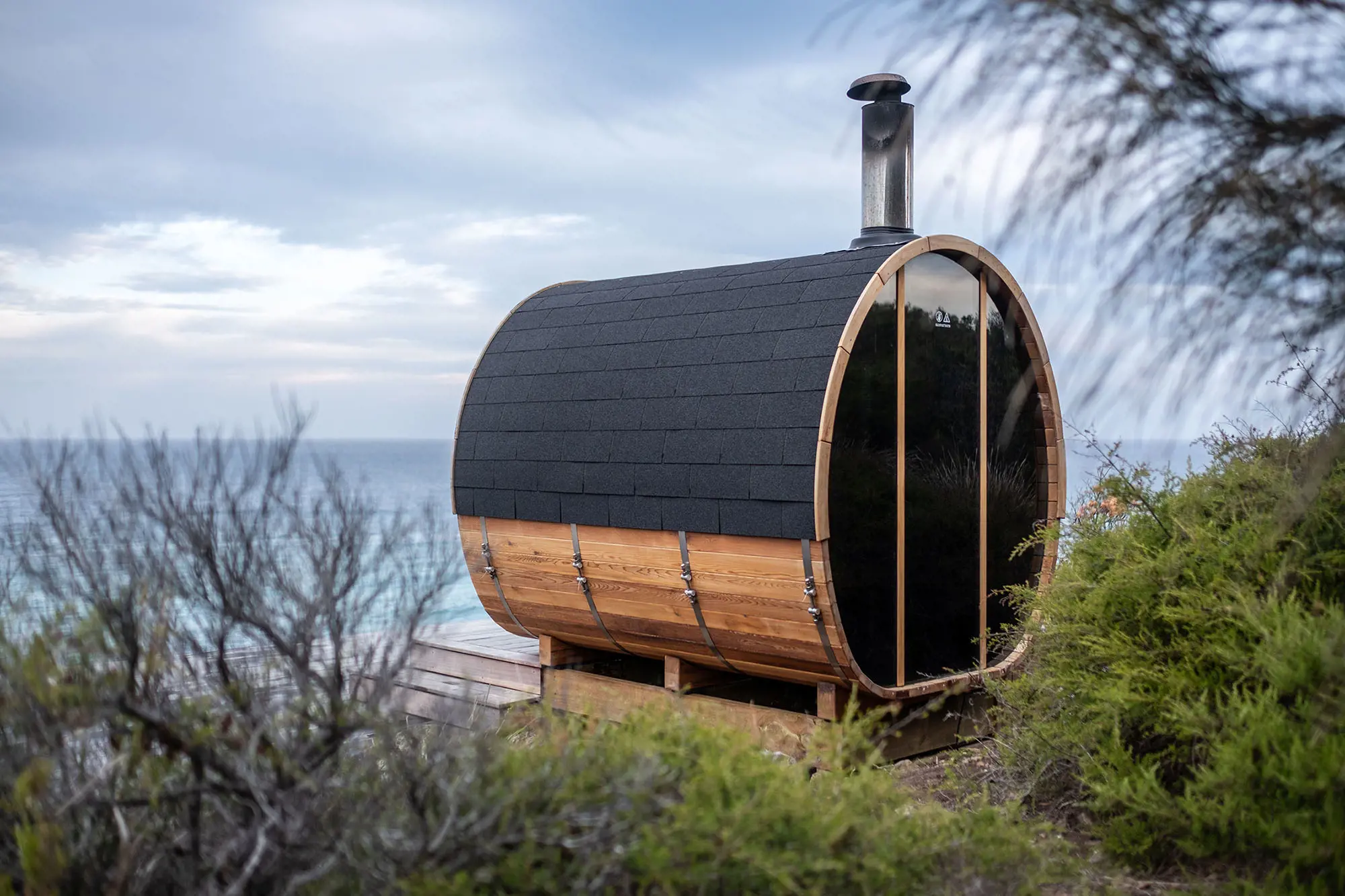 A barrel-shaped wooden sauna with a black shingled roof is situated on a deck overlooking the ocean, surrounded by coastal vegetation.