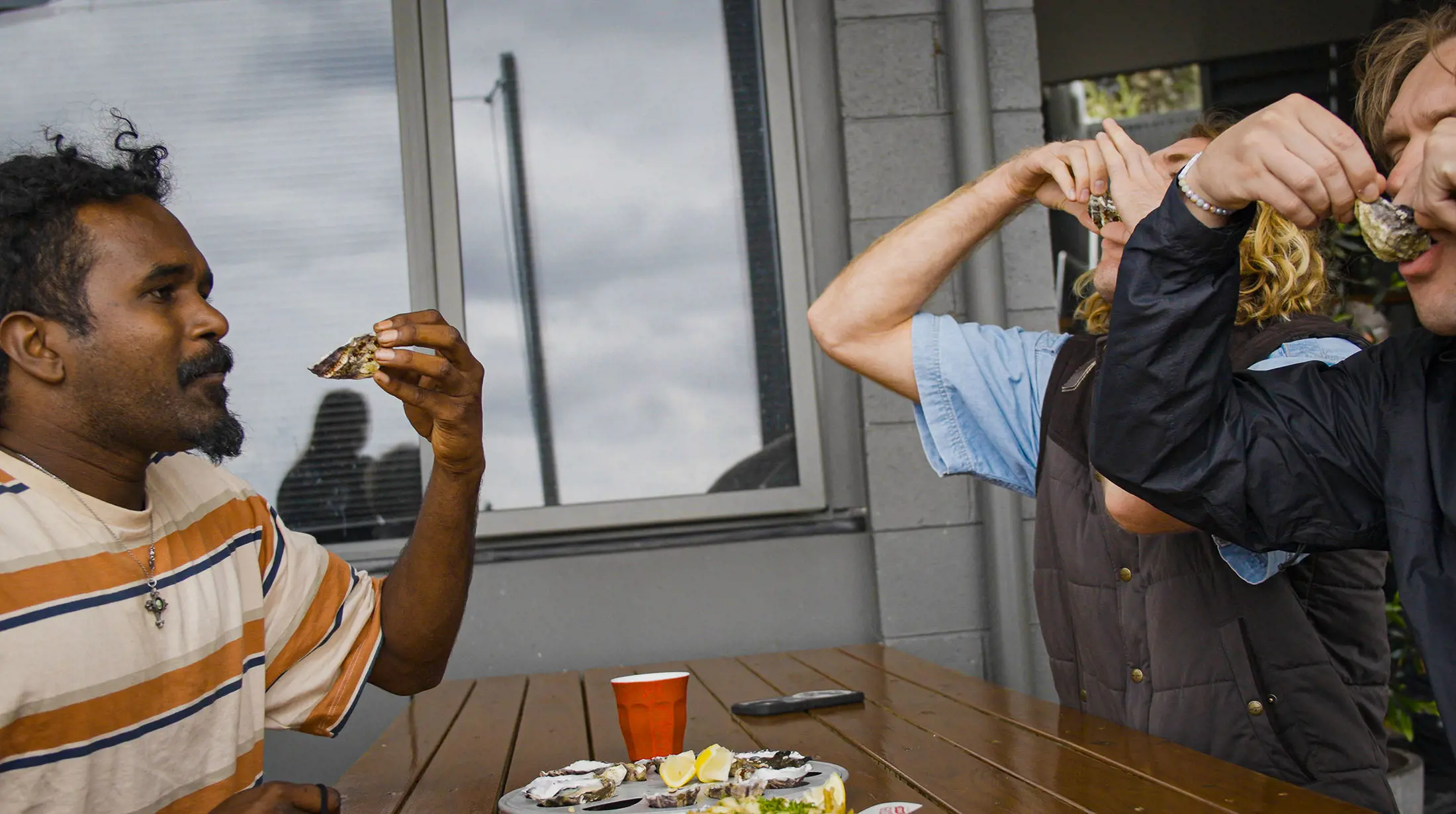Three people sitting at a wooden table, slurping oysters from their shells. A platter of oysters and lemon wedges sits on the table between them.