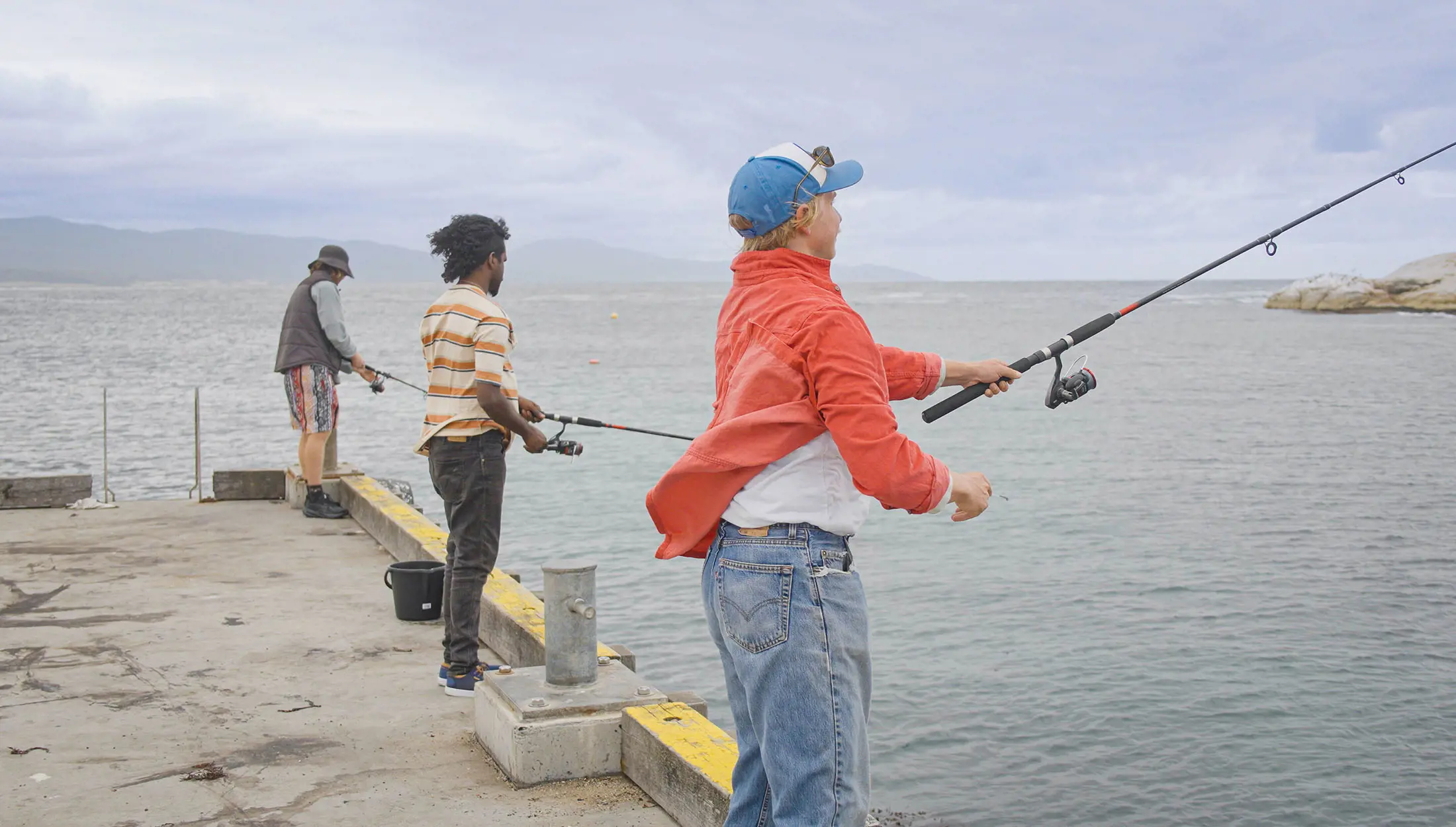 Three people are fishing off a concrete pier by the sea, with mountains visible in the background.