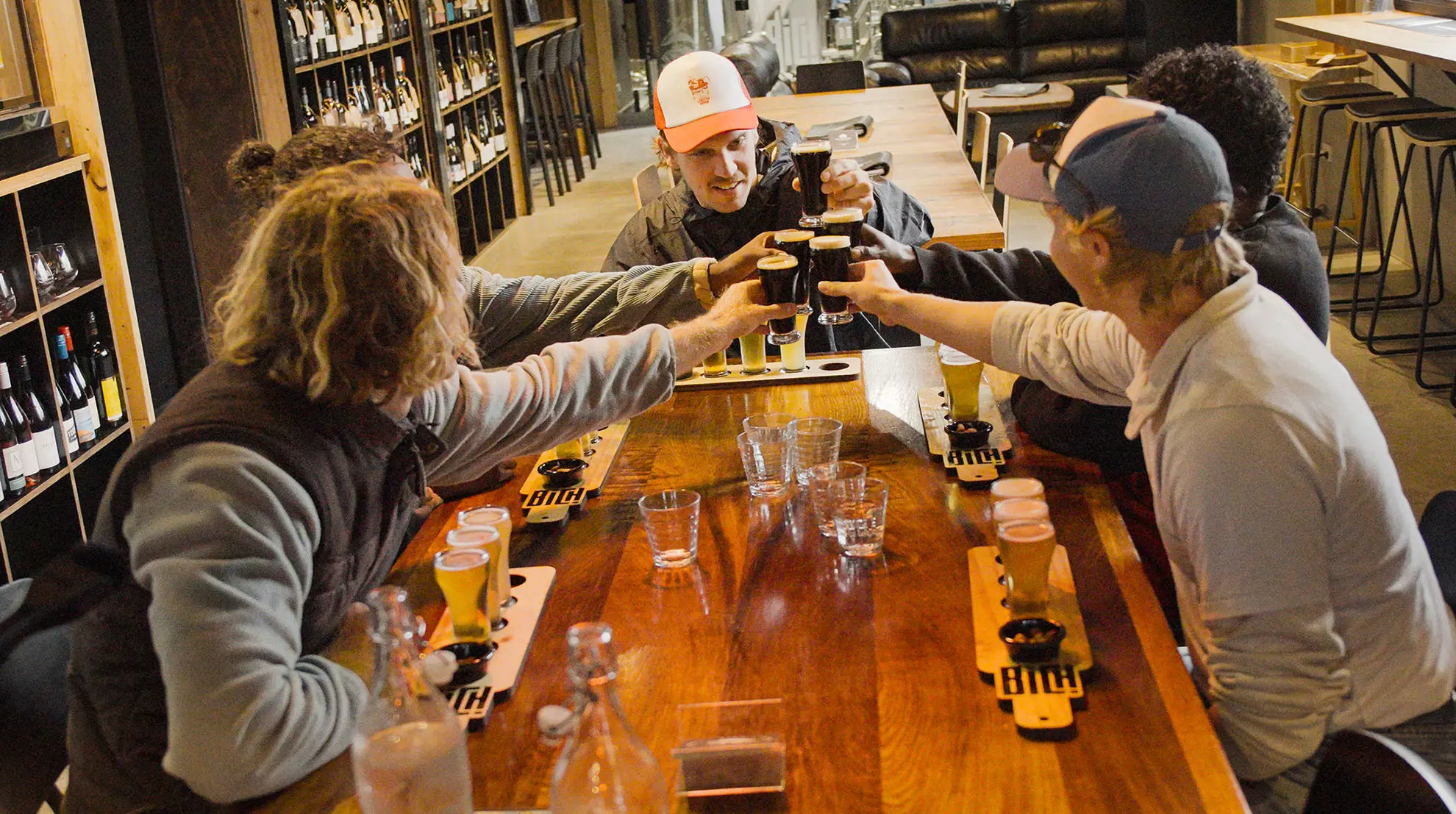 Five people are seated around a wooden table in a pub, raising their glasses for a toast. Each has a tasting paddle of beer in front of them.