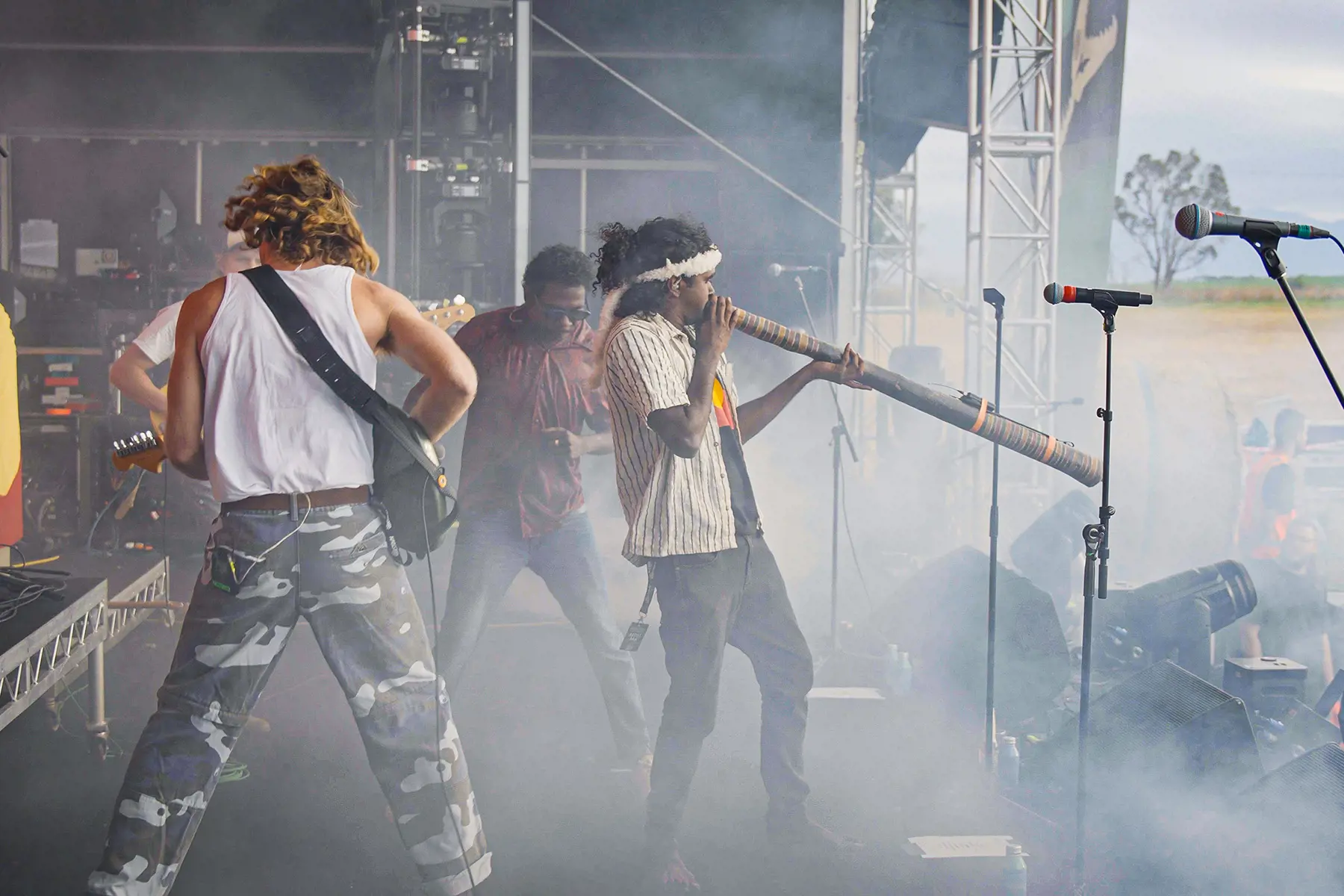 A band performs on a smoky outdoor stage. One musician plays an electric guitar, another plays a yidaki, and other band members are partially visible. Equipment and microphones are set up around them.