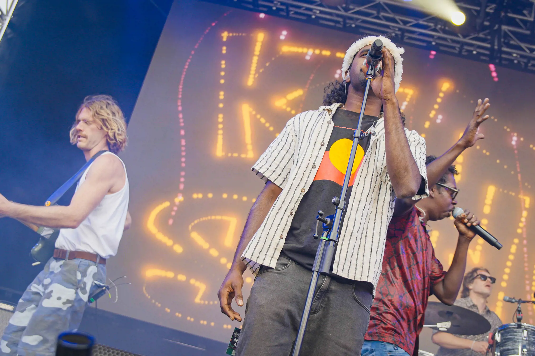 A band performs on stage with microphones and a guitar. The lead singer wears a striped shirt over a t-shirt bearing the Aboriginal flag. The background features bright, illuminated text reading 'King Stingray'.