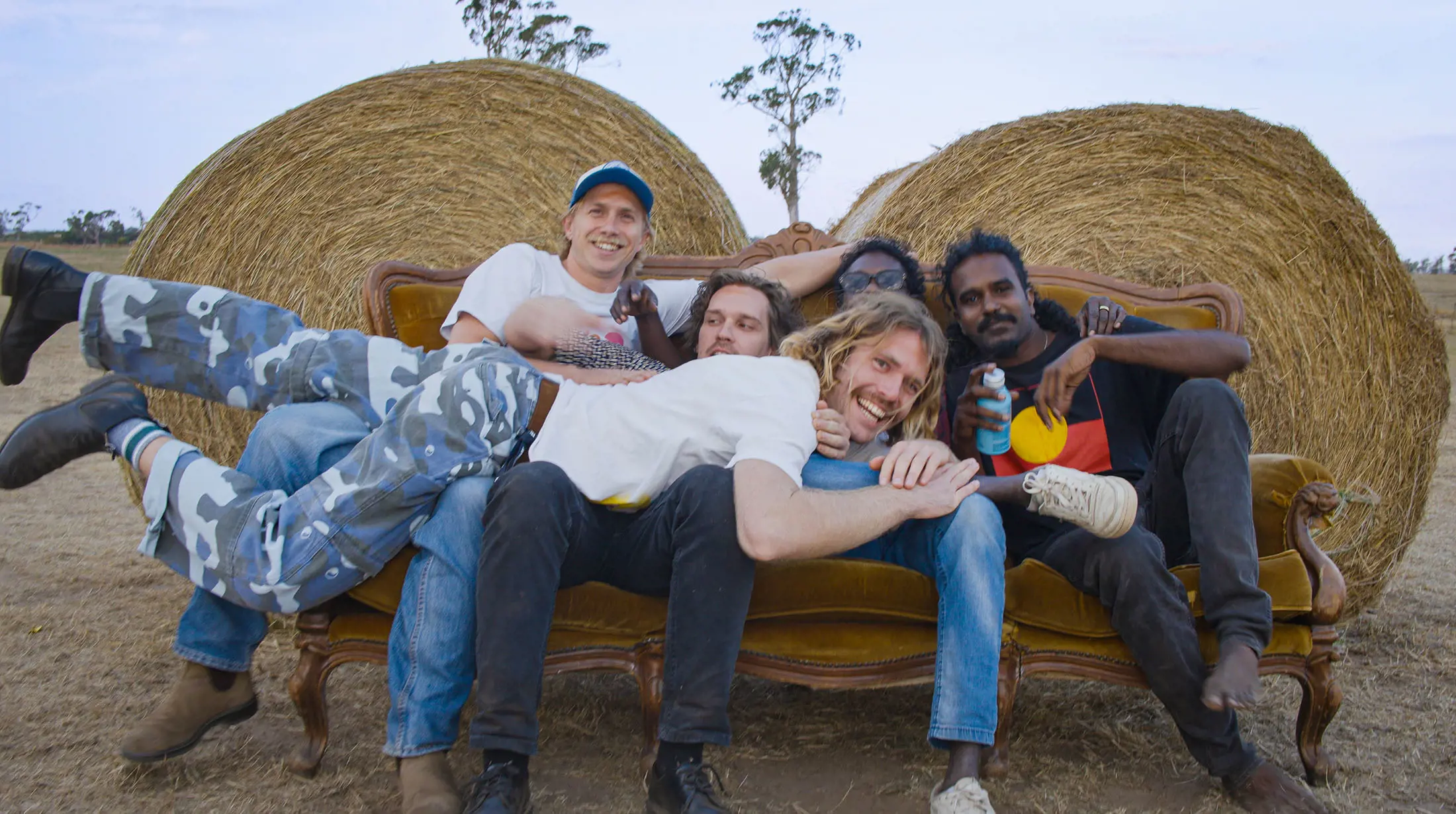 Five people are piled onto a vintage sofa, placed outdoors in front of two large hay bales.They are happy and relaxed, laughing for the camera and holding beers.