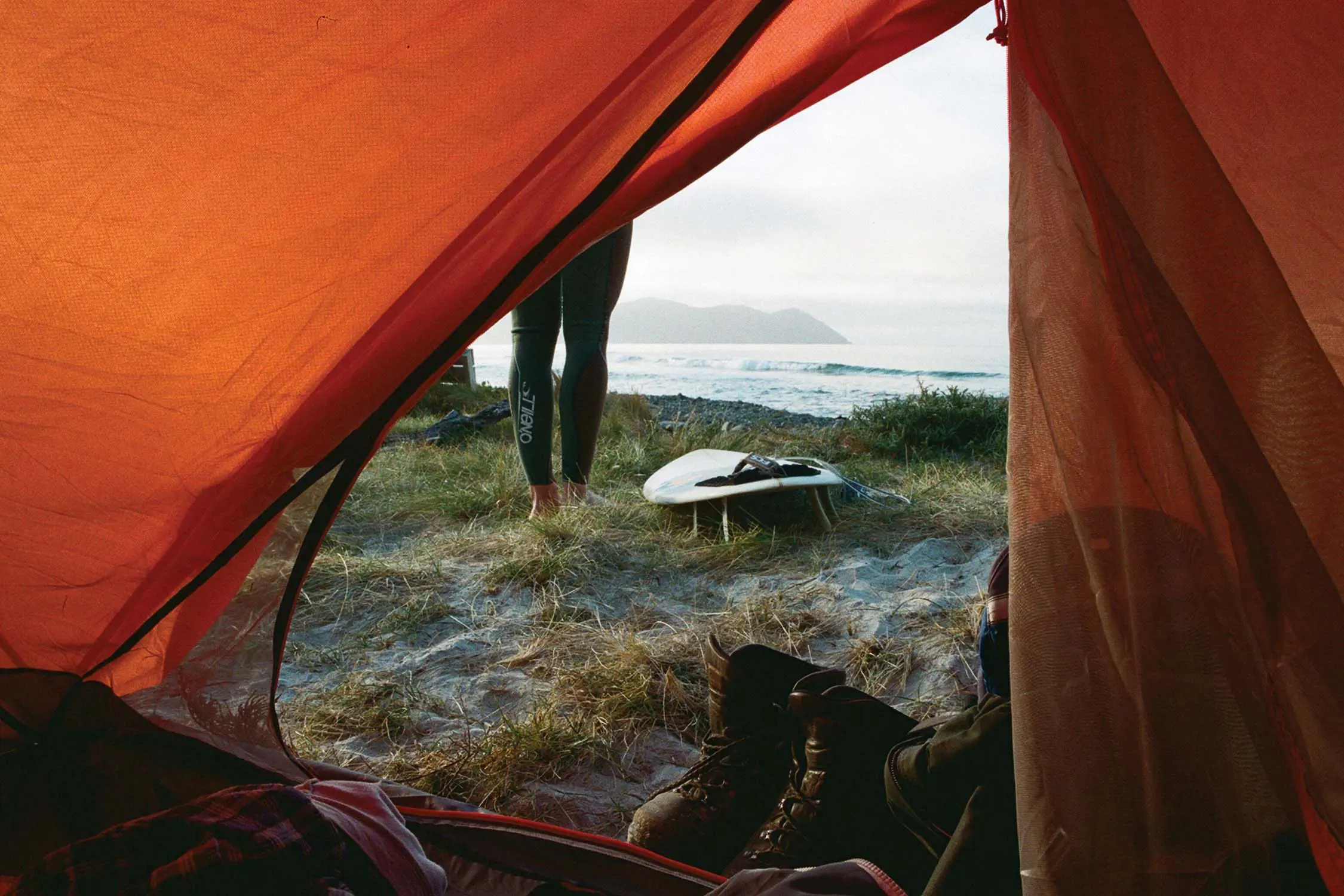 Inside an orange tent, looking through the unzipped door. Outside on the sandy grass, a person's wetsuit-clad legs are seen next to a surfboard.