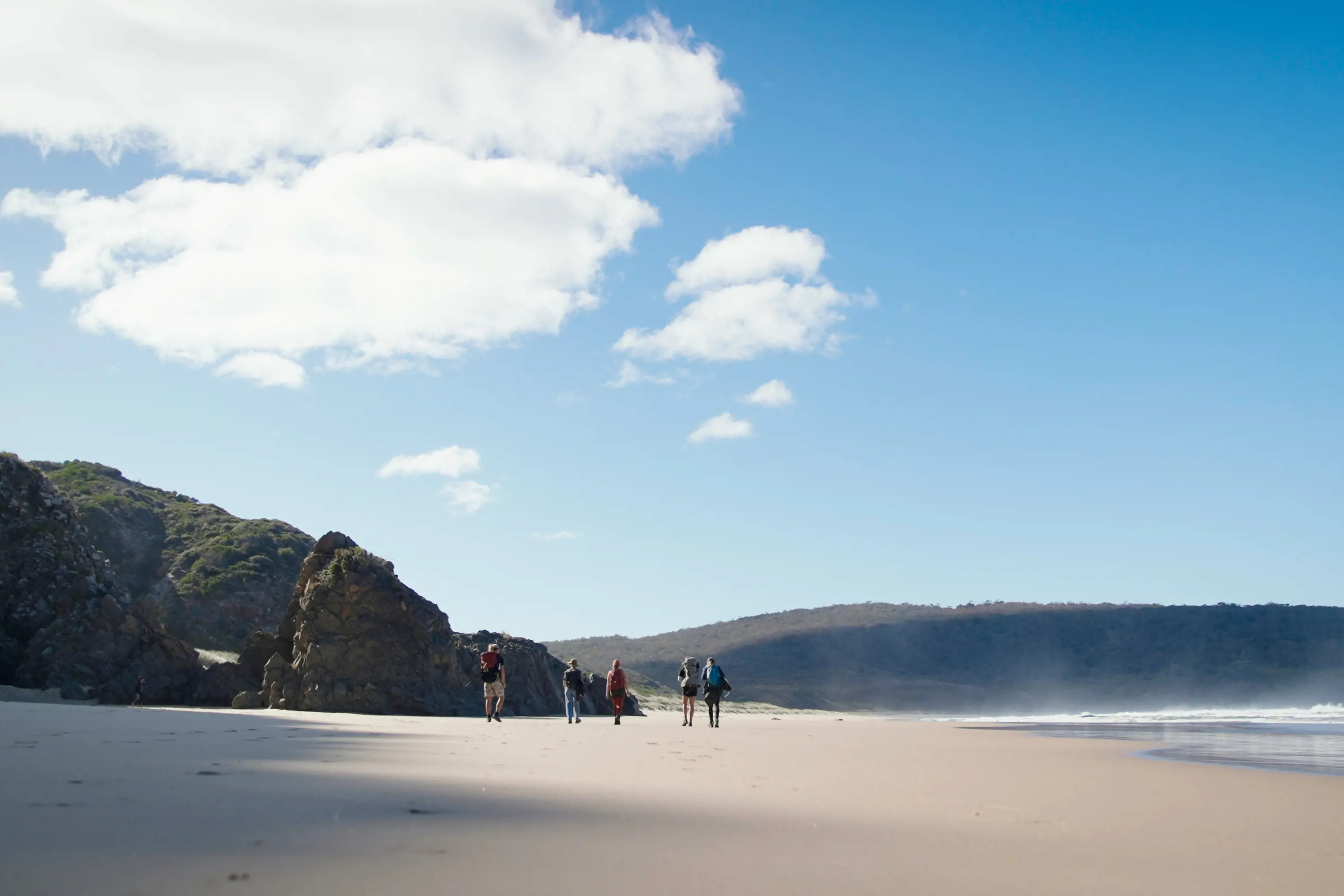 A group of people in the distance walk along a wide beach, with steeply rising bush landscape in the distance.