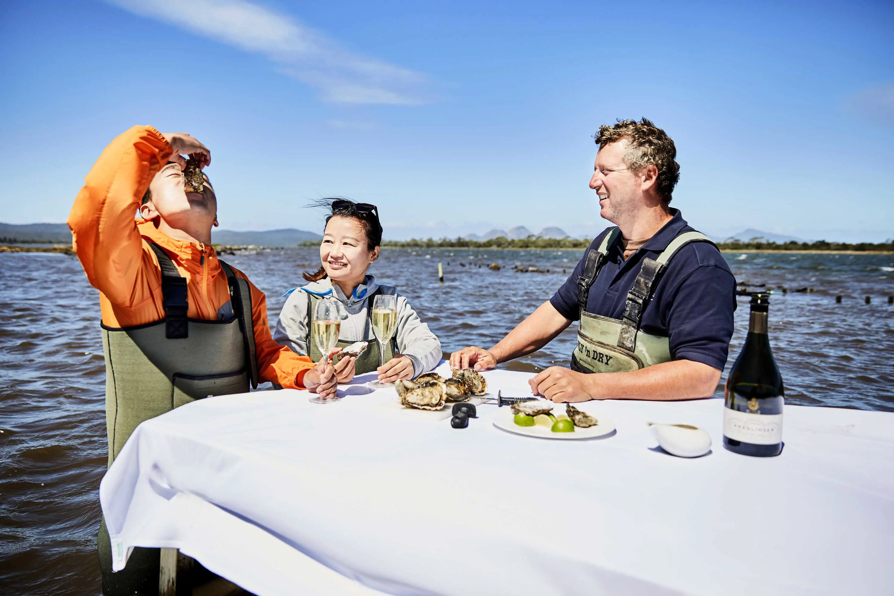 A young couple sit at a table covered in a white table cloth, set up in shallow waters at an oyster farm.