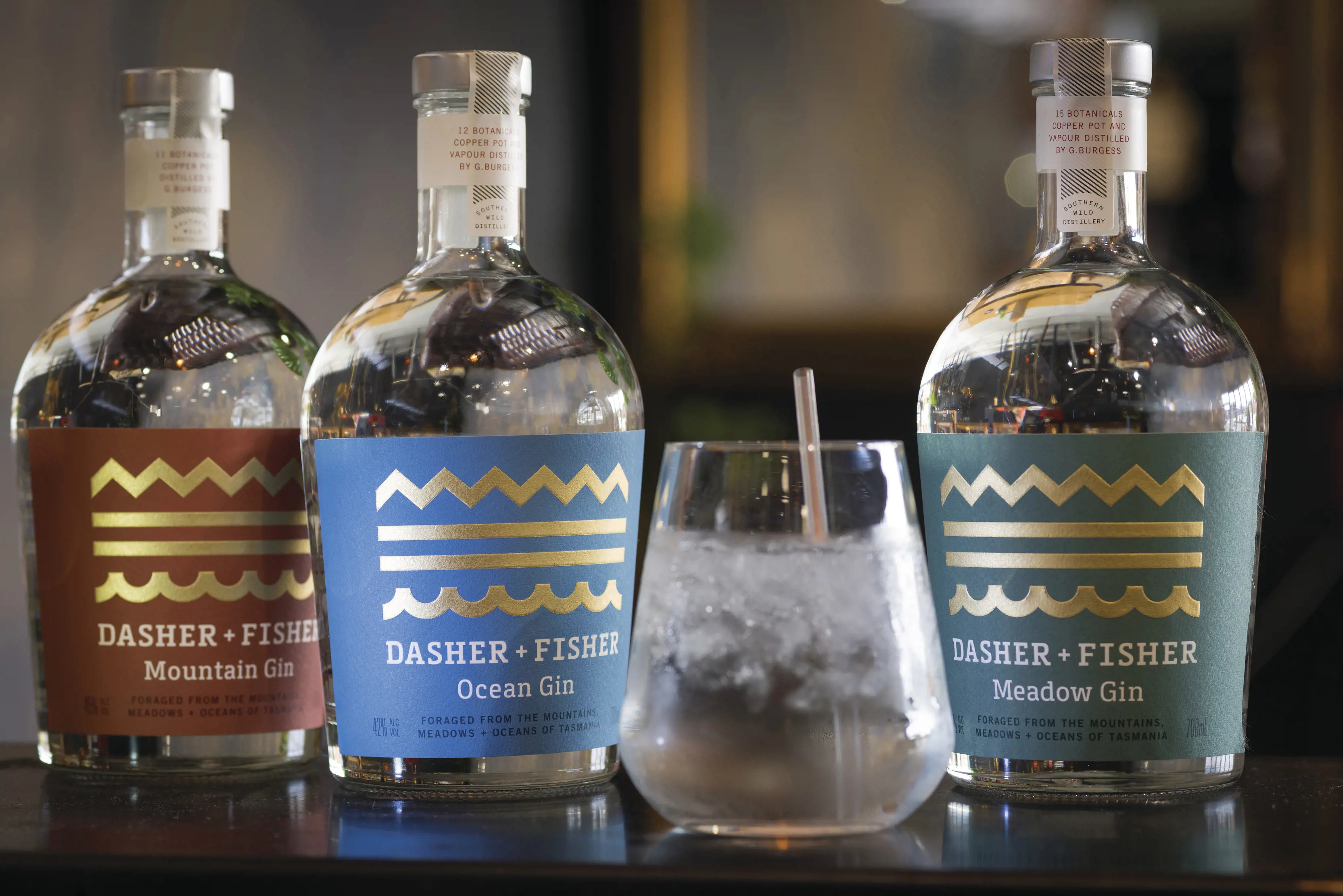Three bottles of Dasher + Fisher Gin and a glass with drink poured at Southern Wild Distillery. The bottle labels include; Ocean, Meadow and Mountain Gin.