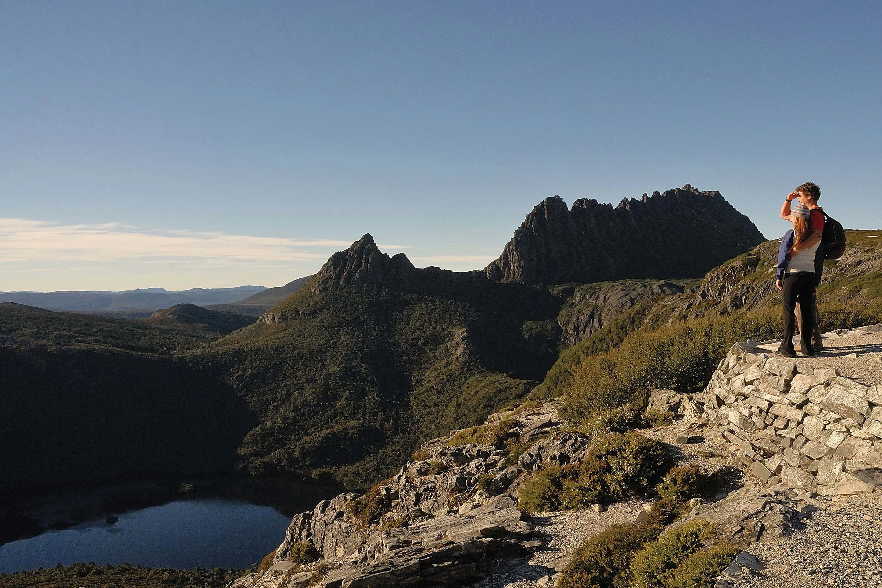 Two people stand on a rough stone wall, looking out over a stunning view of Dove Lake and Cradle Mountain.