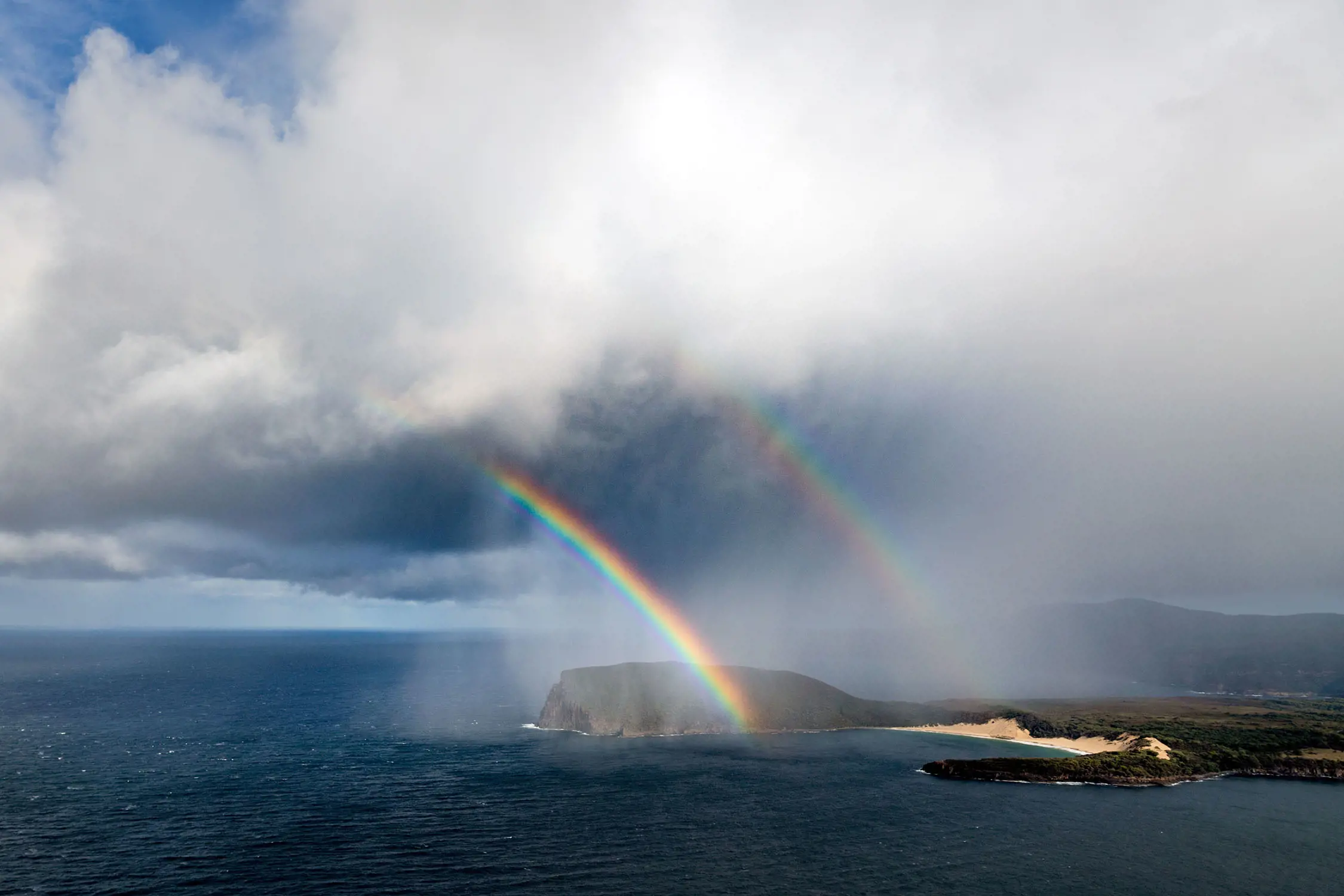 From a lookout point, looking over a peninsula in the distance. Two rainbows are descending to the land, from heavy, low-hanging rainclouds above.
