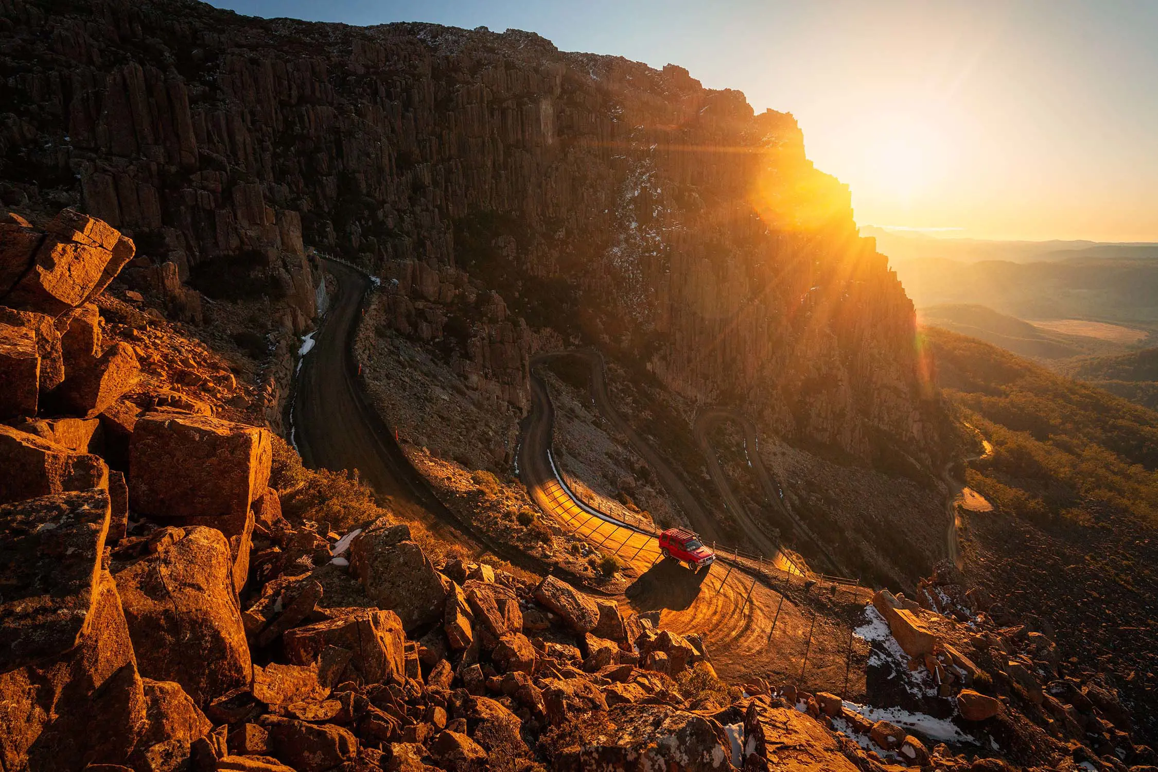 A dramatic rocky cliff illuminated by the setting sun, with a winding dirt road and a parked vehicle at its base. The warm sunlight highlights the rugged terrain and creates long shadows, emphasizing the texture of the rocks.