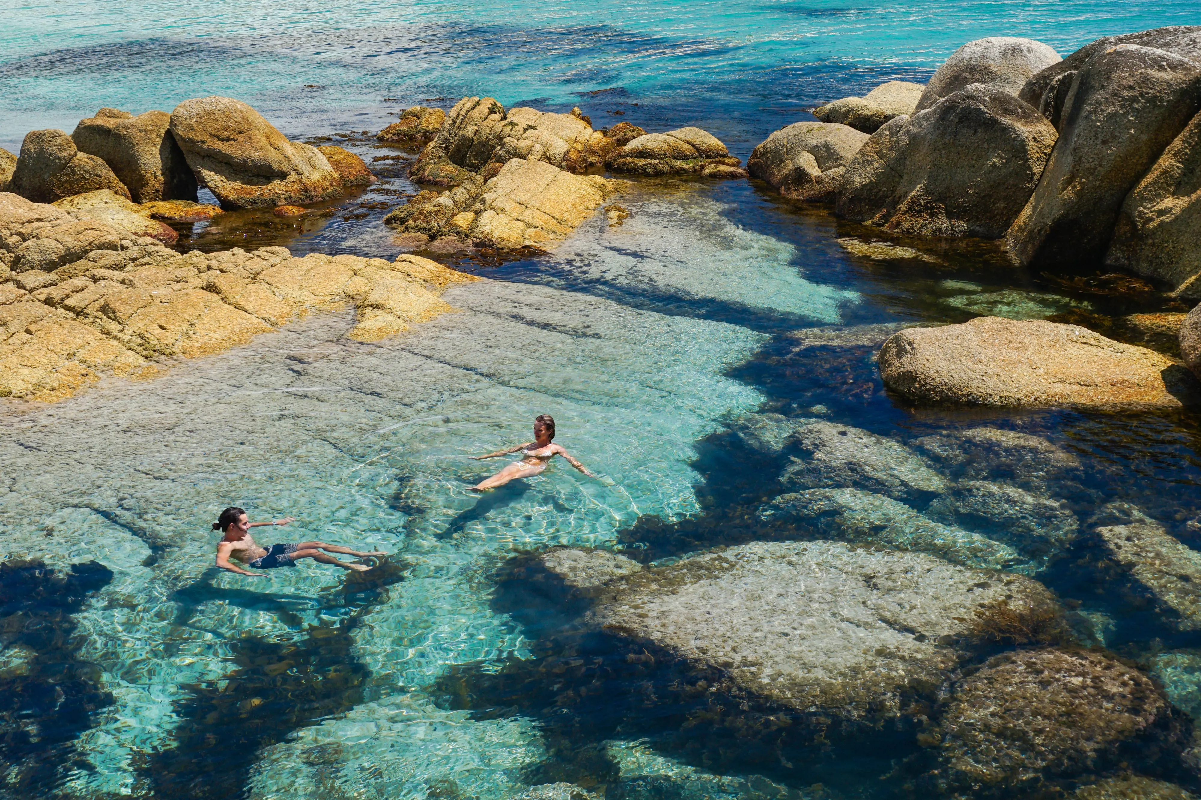 Two swimmers float in the crystal clear, light blue waters of a sheltered rockpool inlet on a sunny day.