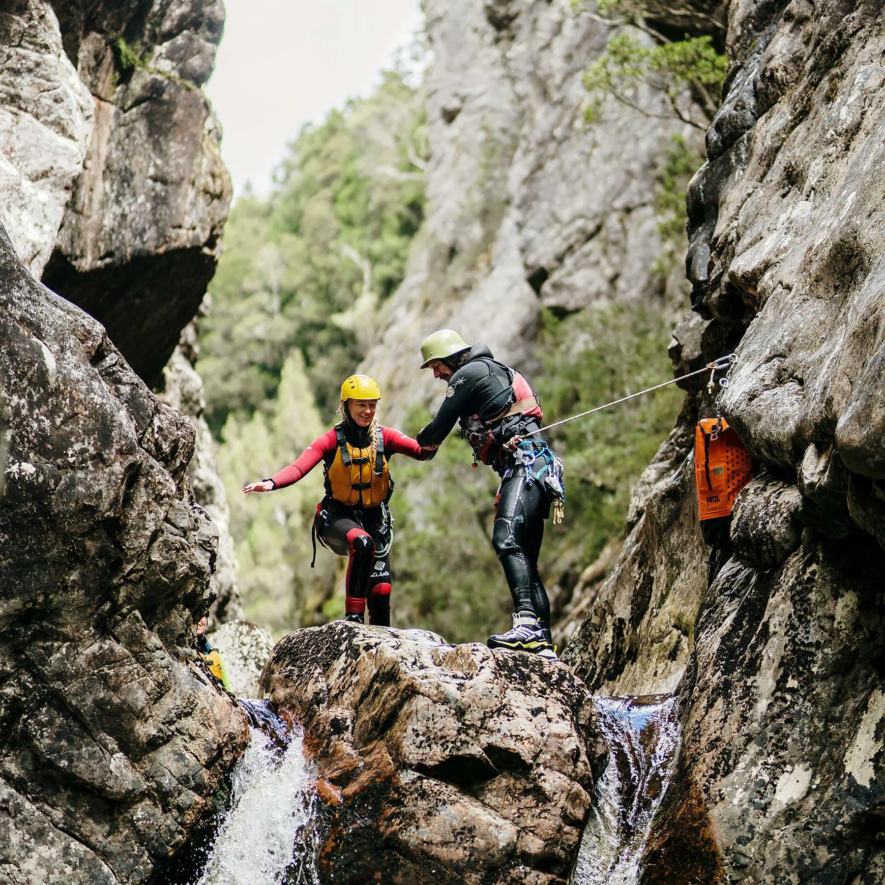 Two people in outdoor water gear, lifejackets and helmets are climbing through a narrow gap in the cliffs where water is pouring through.