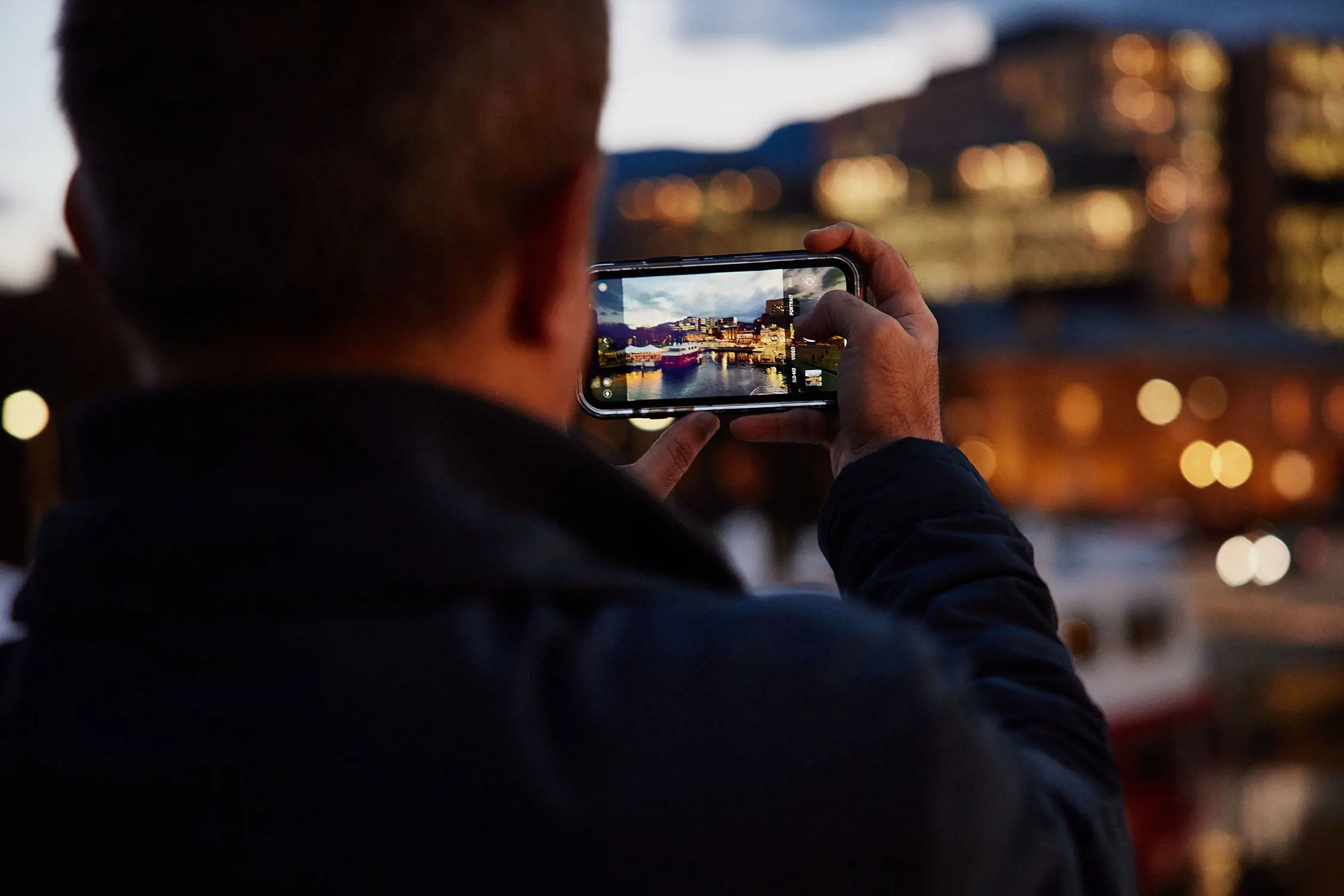 Looking over a person's shoulder as they hold their phone up to take a photo of the Hobart waterfront area at dusk.