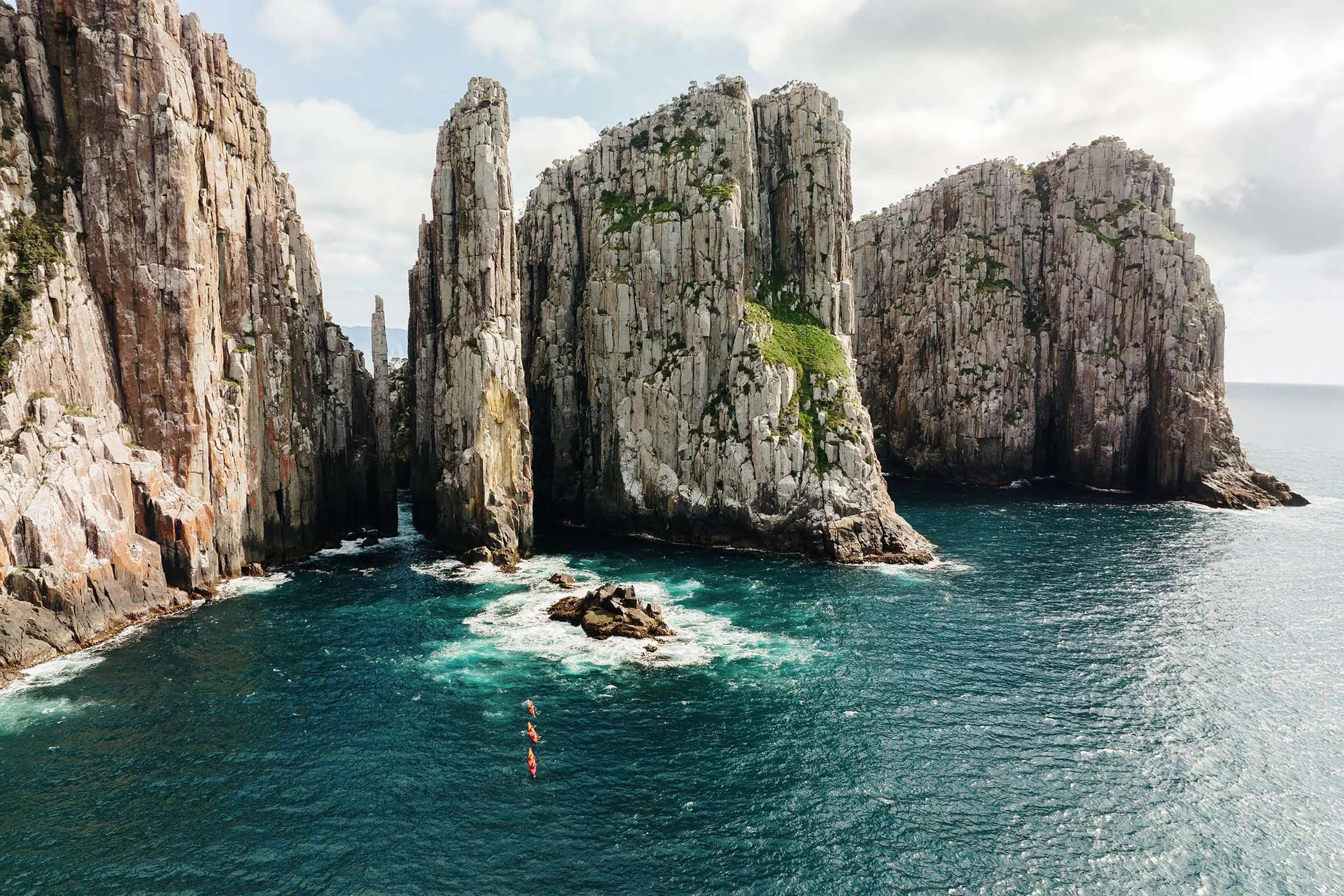 An aerial view of three kayaks in a line in the dark blue ocean waters, dwarfed by some sharply rising cliffs that tower above them.