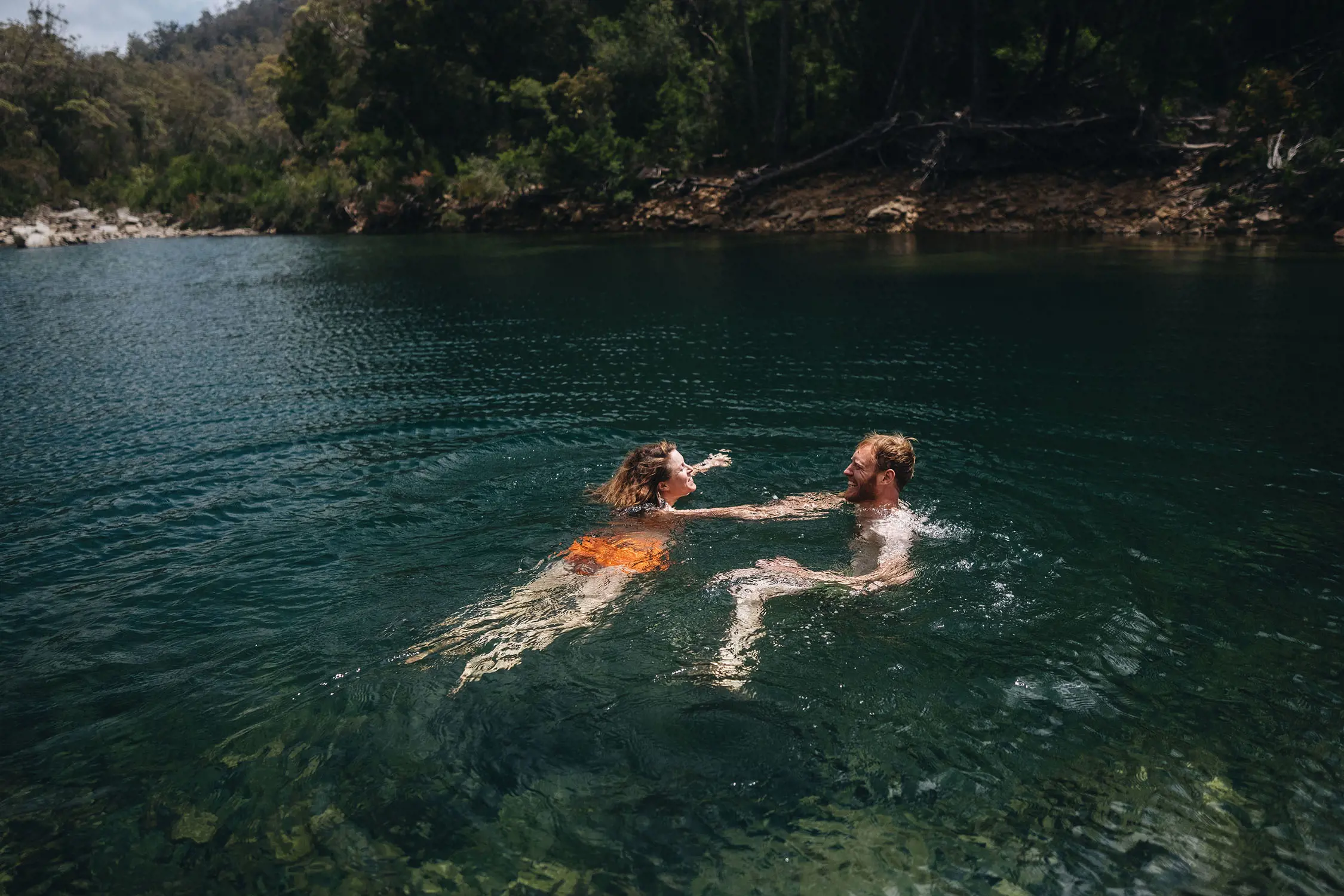 Two swimmers in the dark green-blue waters of a lake. The thick bush surrounds the banks of the watering hole.