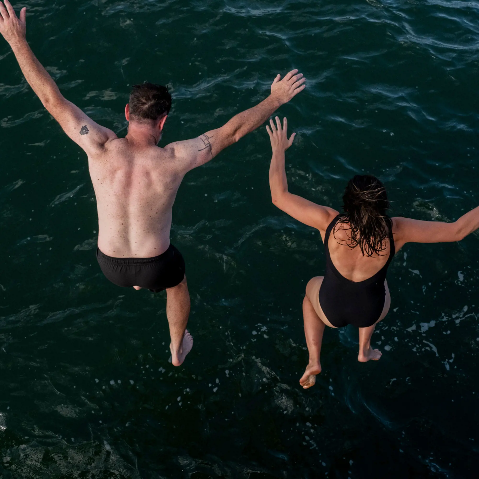 Two people in swimming gear jump, with arms outstretched, into the dark water of a lake