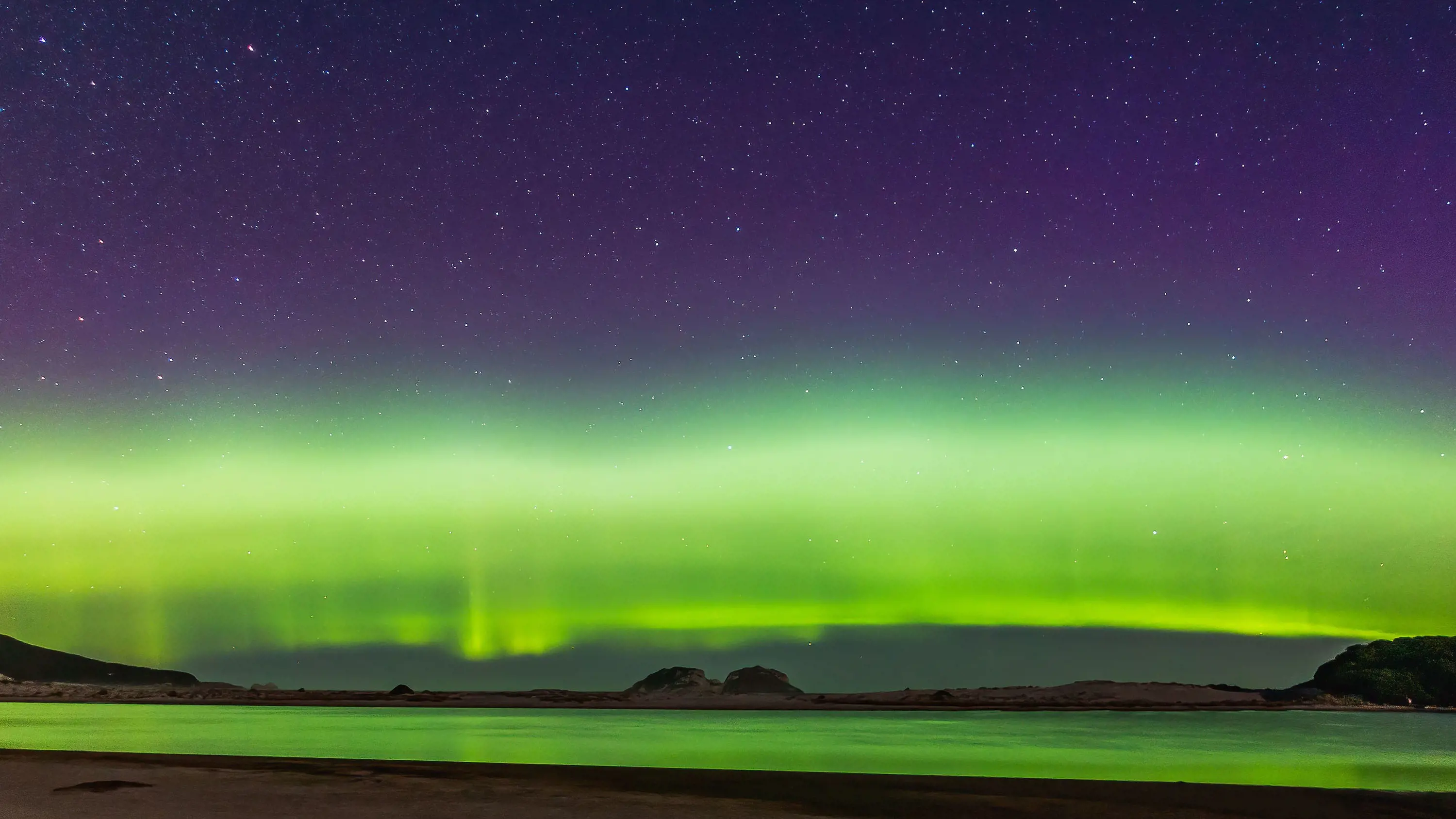 Standing on a beach at night, bright bands of green aurora hug the horizon and reflect into the water. The sky appears rich, dark and full of stars above it.
