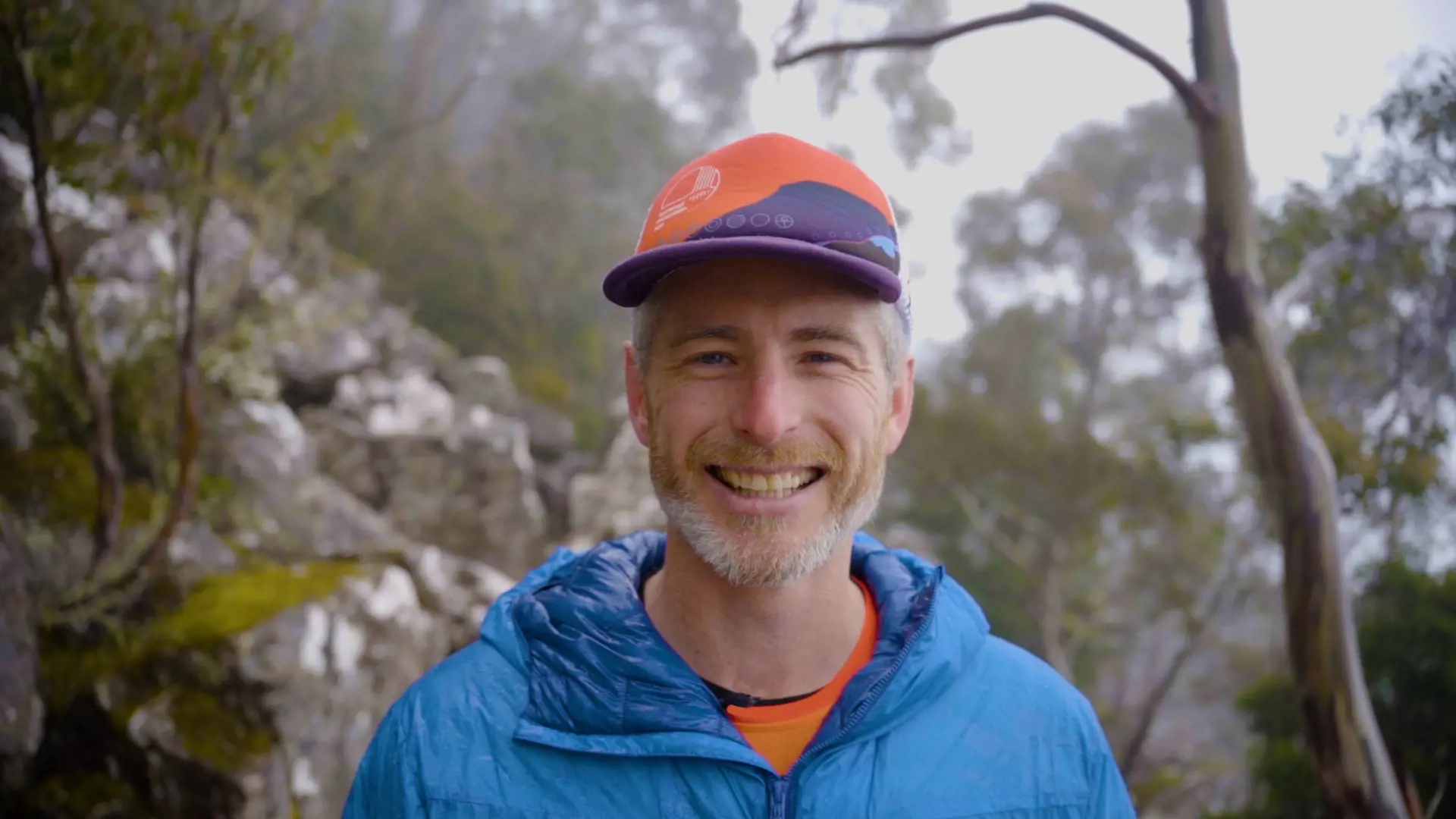 A head and shoulders shot of a smiling man standing in the bush, dressed in a blue puffer jacket and orange and purple cap.
