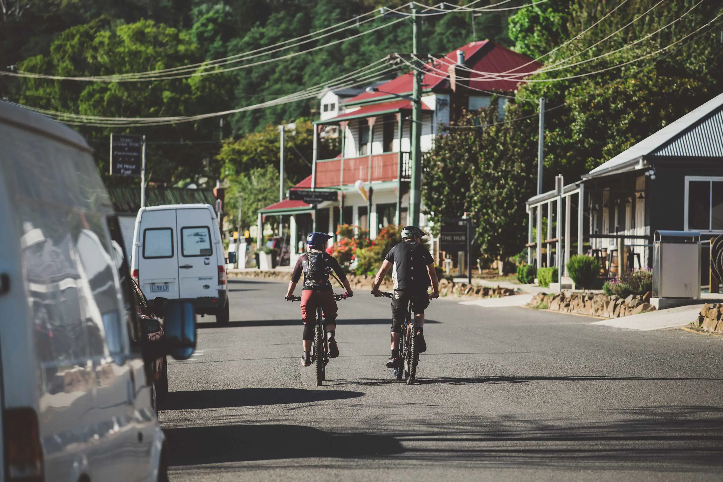 Two mountain bike riders ride their bikes along bitumen throuhg a country town.