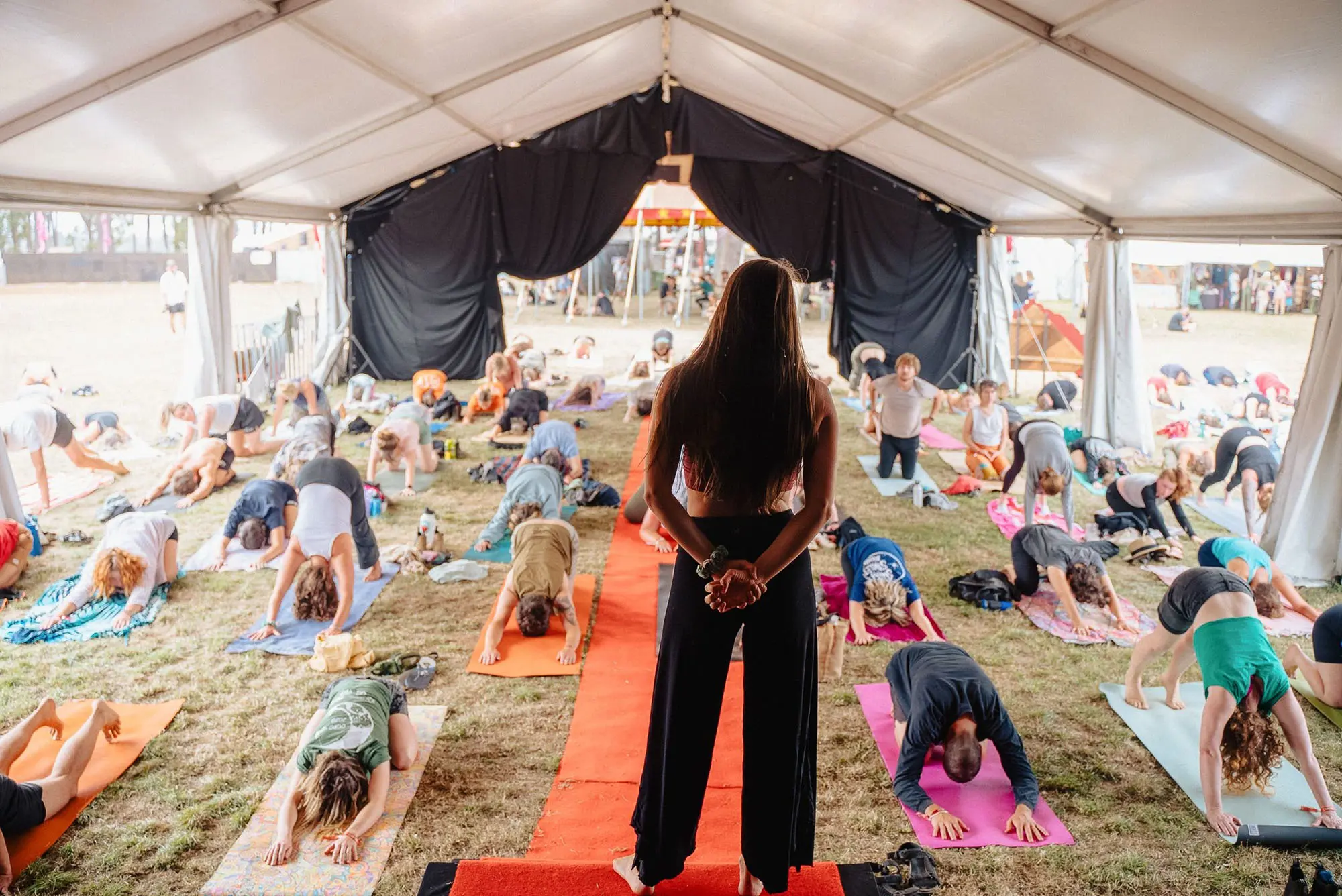 A large group of people on yoga mats are spread out on the grass inside a large marquee tent. They are all mid-pose, mostly facing downwards. A woman stands at the front on a raised platform, leading the class.