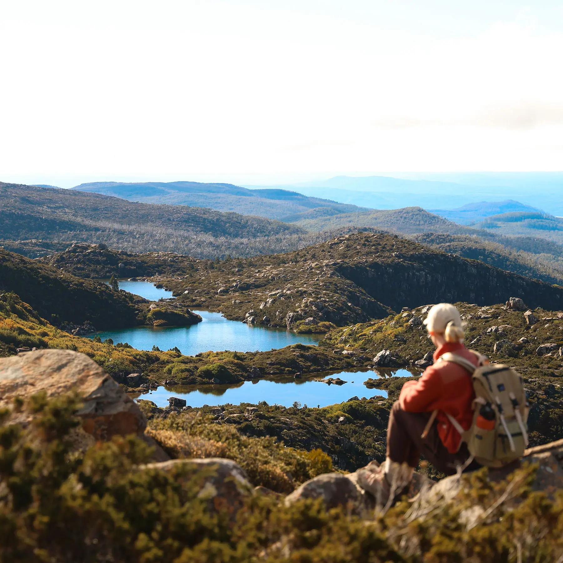 A hiker sits atop a rock, gazing out at the alpine landscape filled with a scattering of blue lakes and rising hills.
