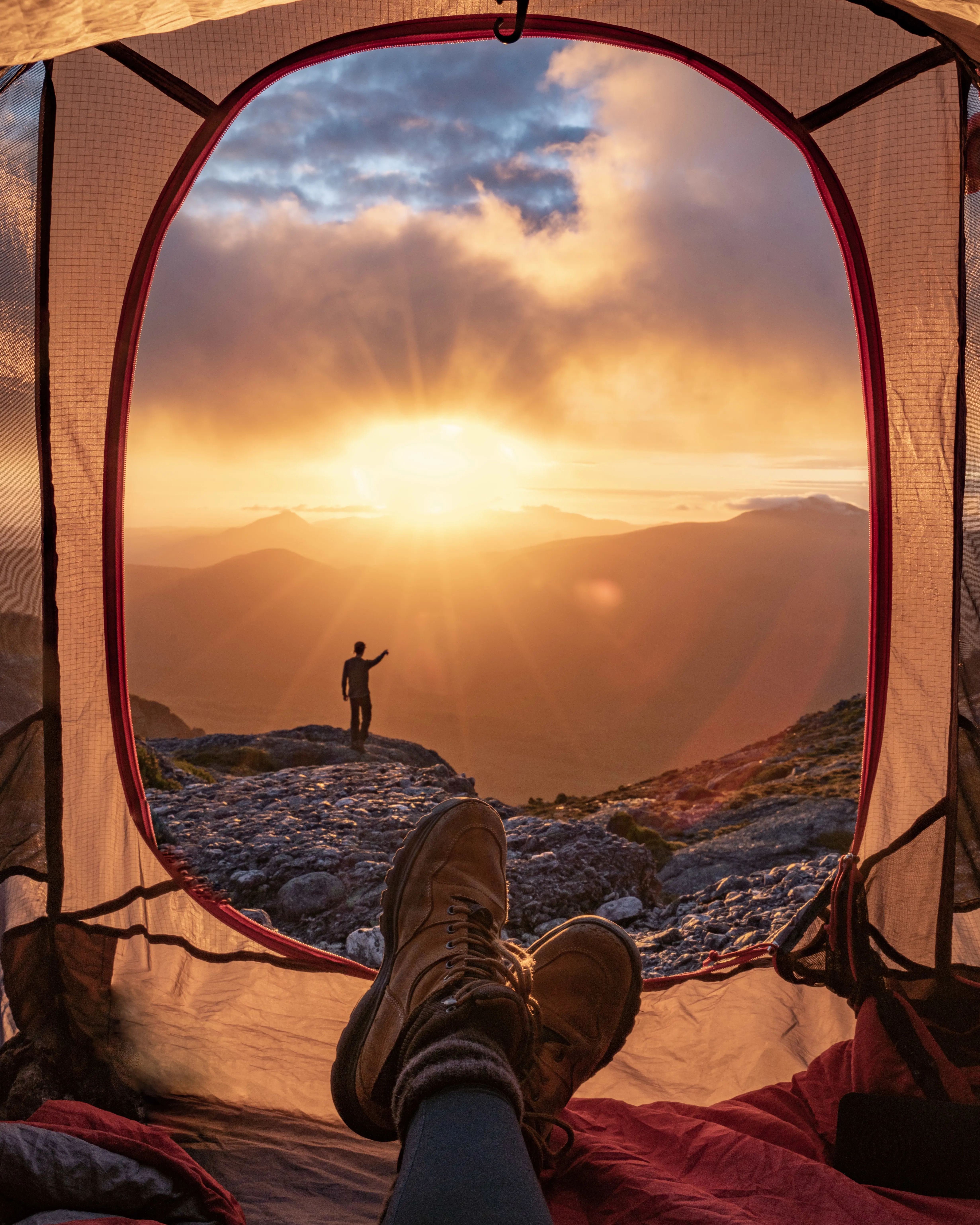 View from inside a tent, with a person outside the tent entrance silhouetted by the stunning setting sun shining between clouds and mountains.