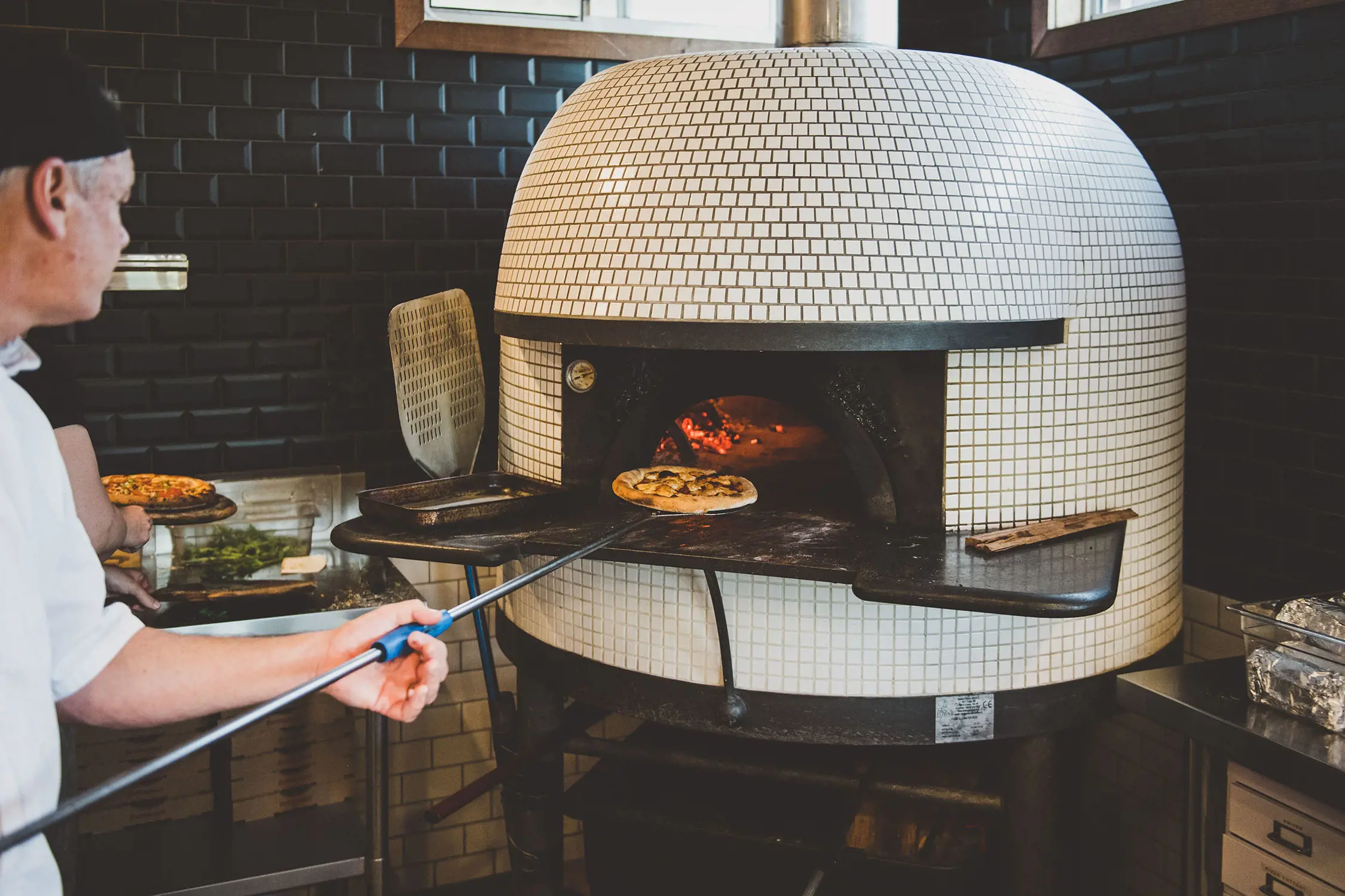 A chef uses a long-handled paddle to pull a cooked pizza from a domed, white-tiled woodfired pizza oven.