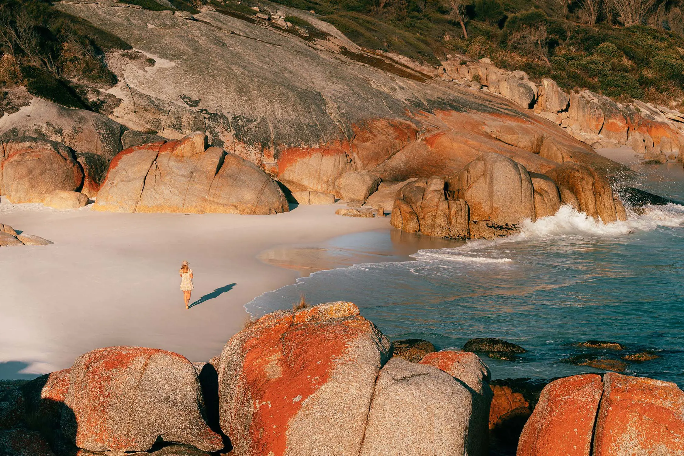 A woman wearing a broad-brimmed hat walks along the sand in a bay sheltered by red-lichen-covered rocks.