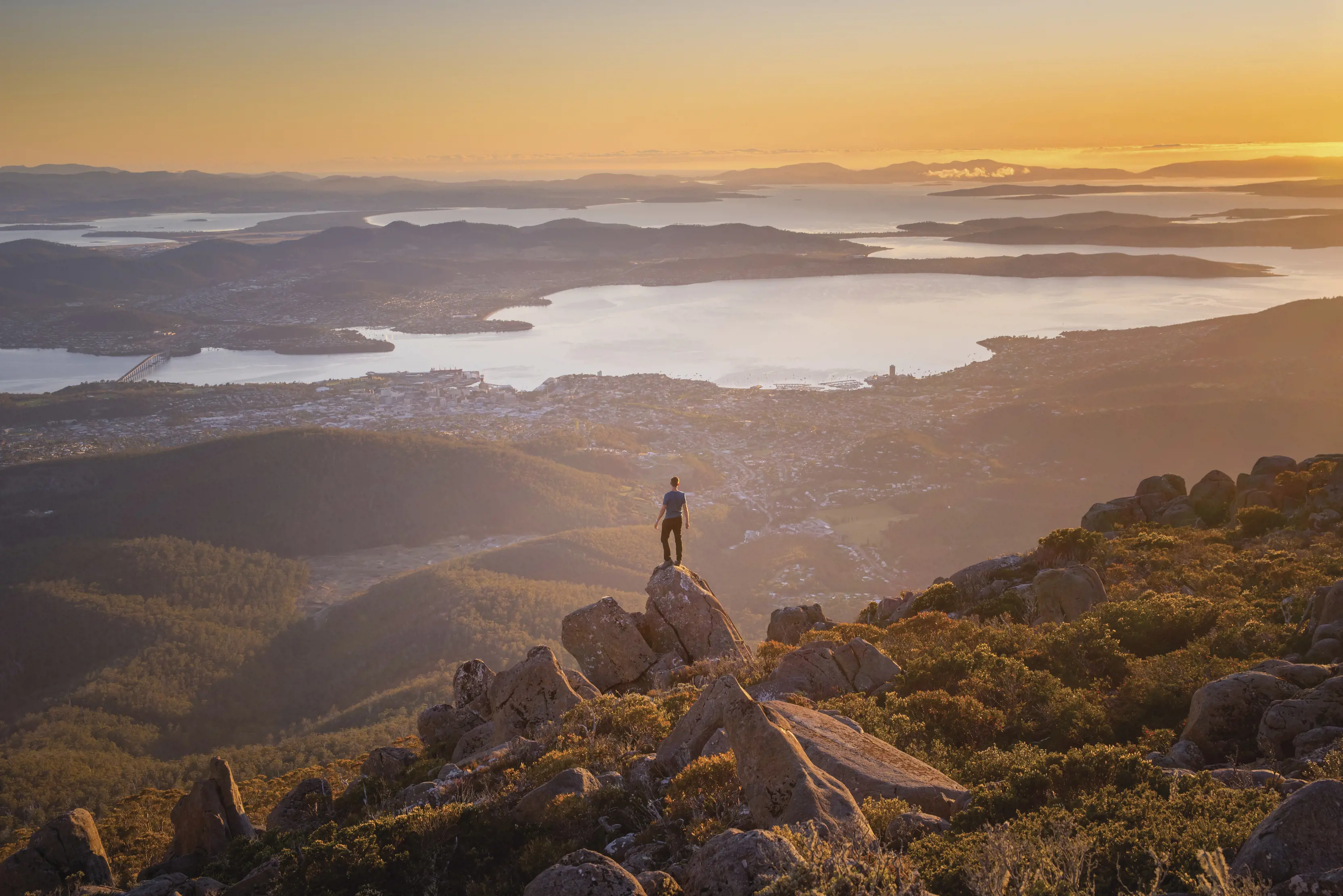 A person stands atop a rocky outcrop on a mountain, overlooking a beautiful scenic cityscape with a river snaking through. The sky is the orange of sunrise.