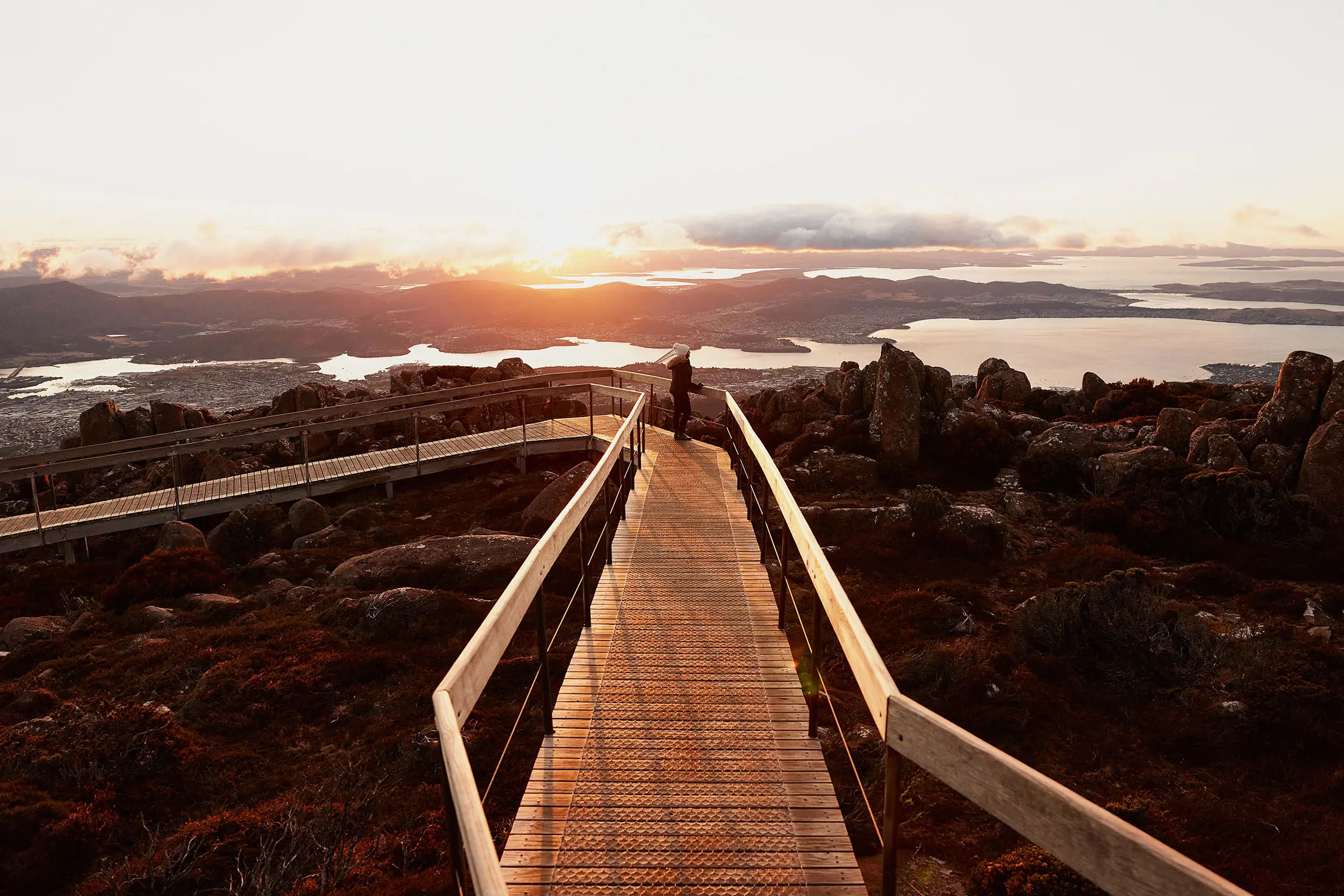 The rock-strewn ground of the summit area with a long wooden boardwalk, and the low-lying sun reflecting off the clouds in the distance.