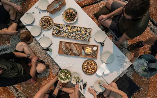 A top-down view of a group around a picnic table perched on an orange-tinged rock. The table is full of oysters, fish and other plates and drinks.