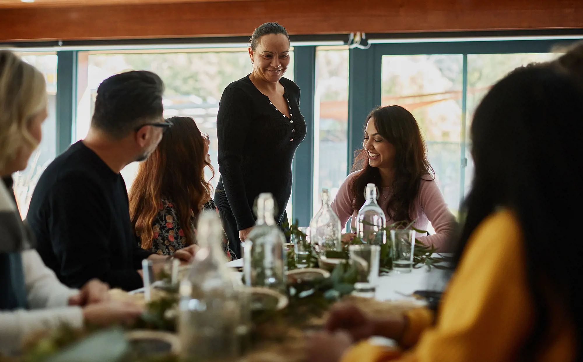 A woman stands next to people seated around a long table, set with foliage, glasses and and water jugs, ready for a meal.