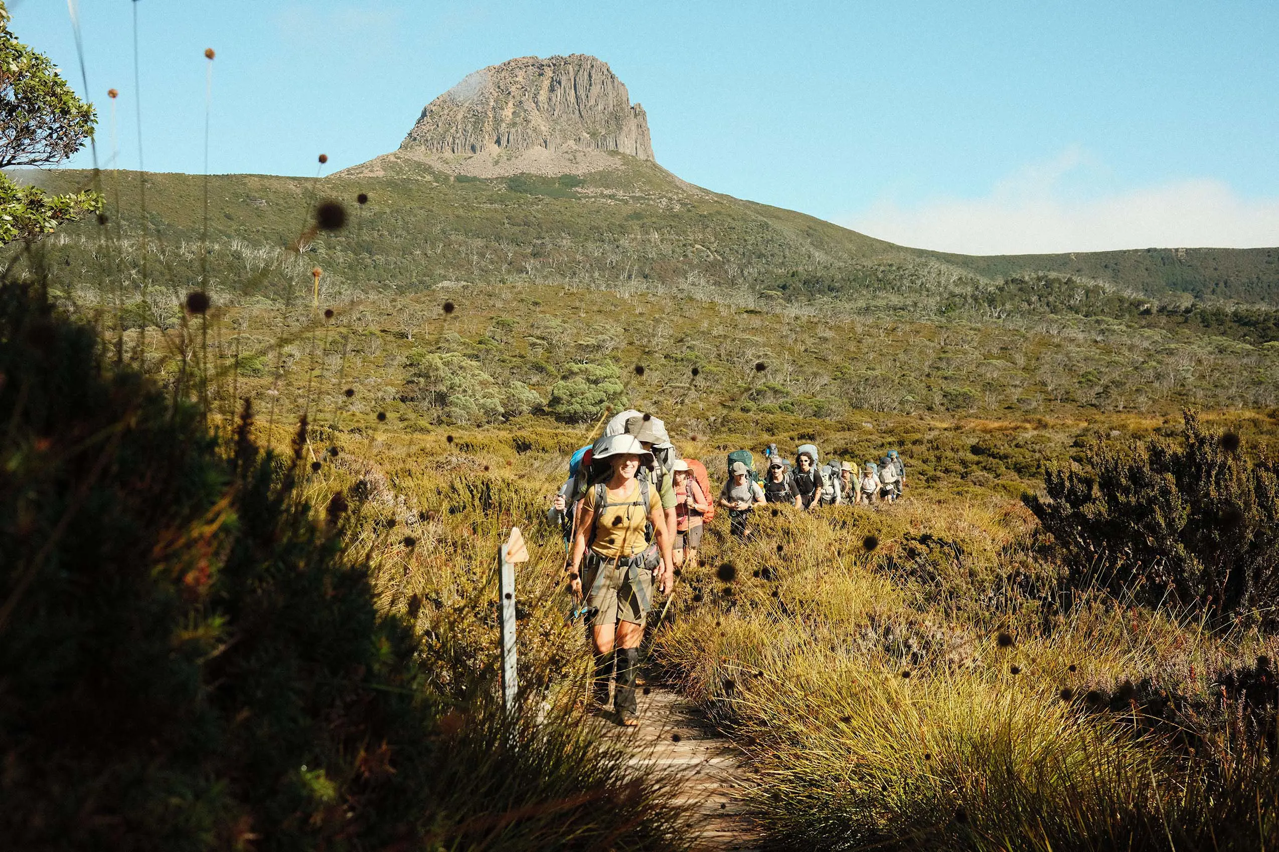 A group of people with hiking gear and large backpacks walk single-file along a trail through the wide-open scrub. Above them, a large mountain peak rises above the horizon.