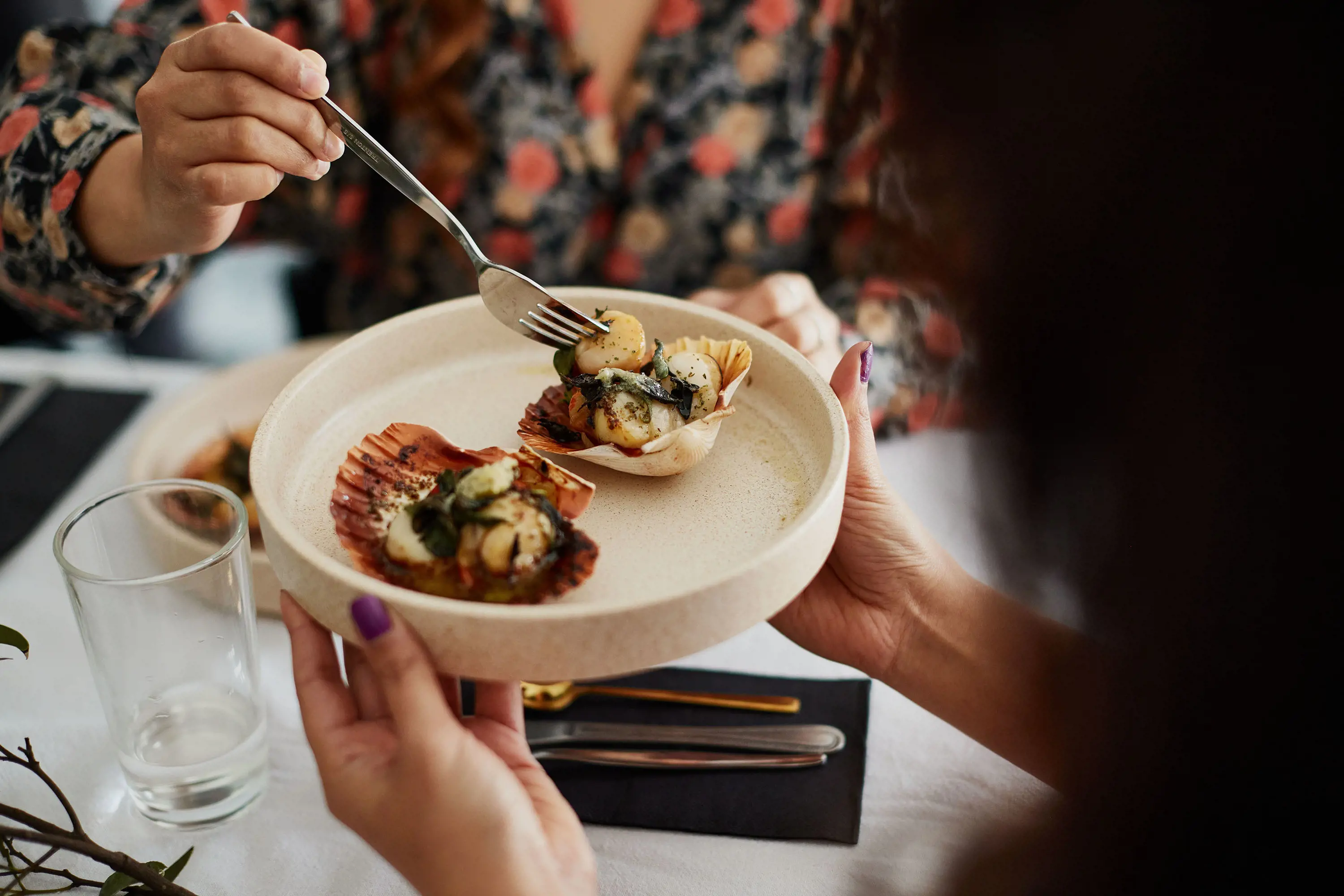 Two delicately plated scallops in their shells are served above a table set for a meal, with one person reaching with their fork to take a scallop.