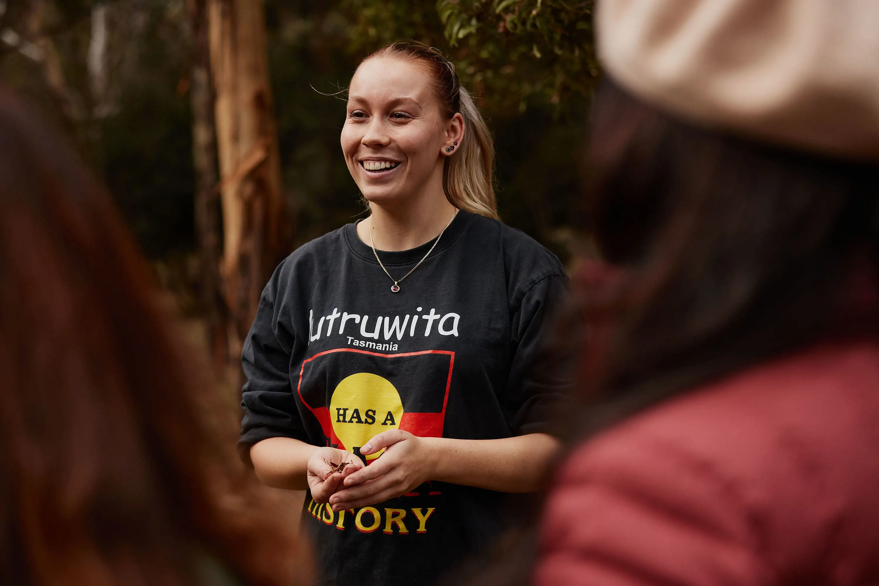 A young woman talks with guests standing in the shade of a tree. Her black shirt features the Aboriginal flag and says "lutruwita Tasmania has a black history".