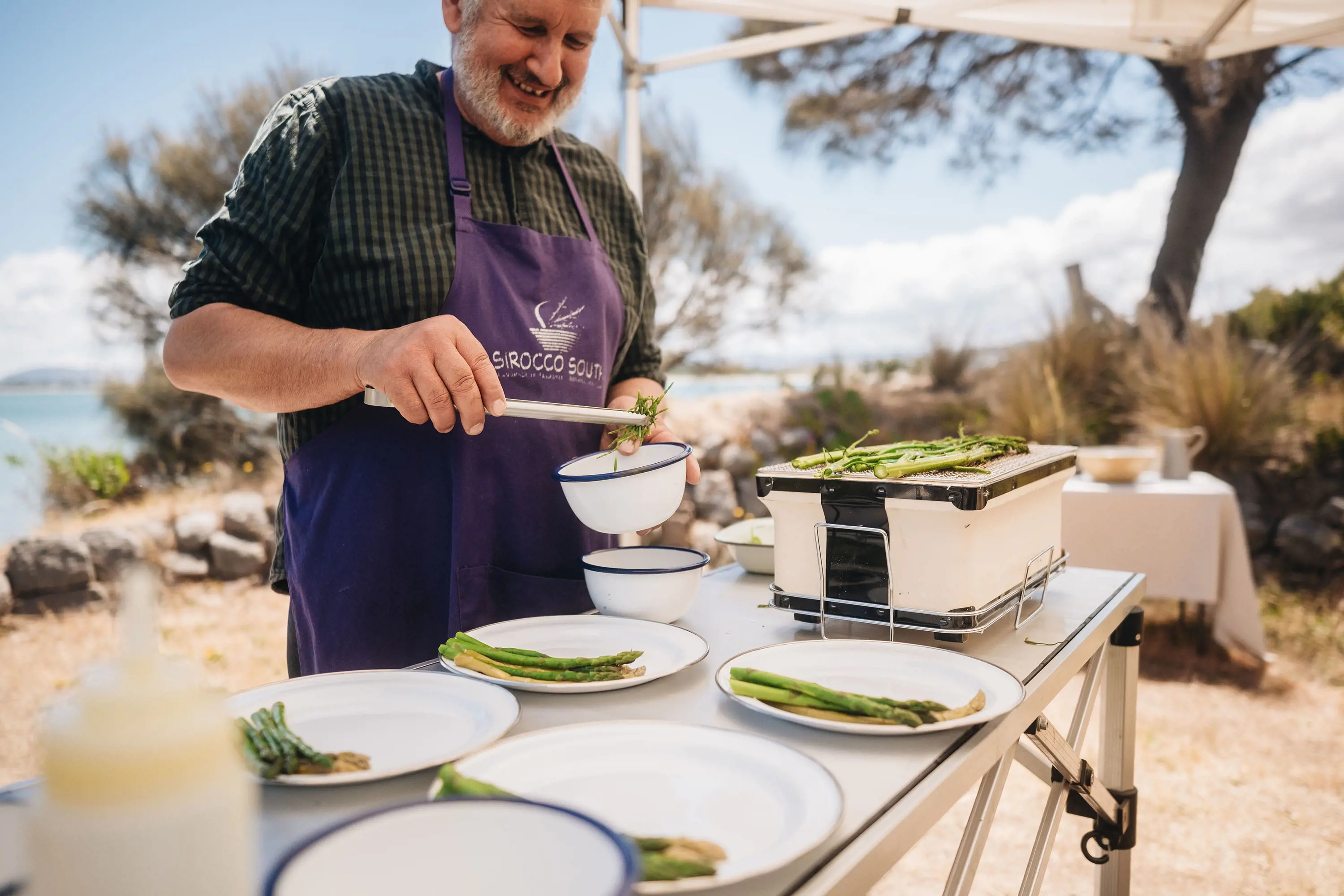 A man wearing a purple apron smiles as he plates up asparagus, cooking on an outdoor table under the shade of a gazebo.