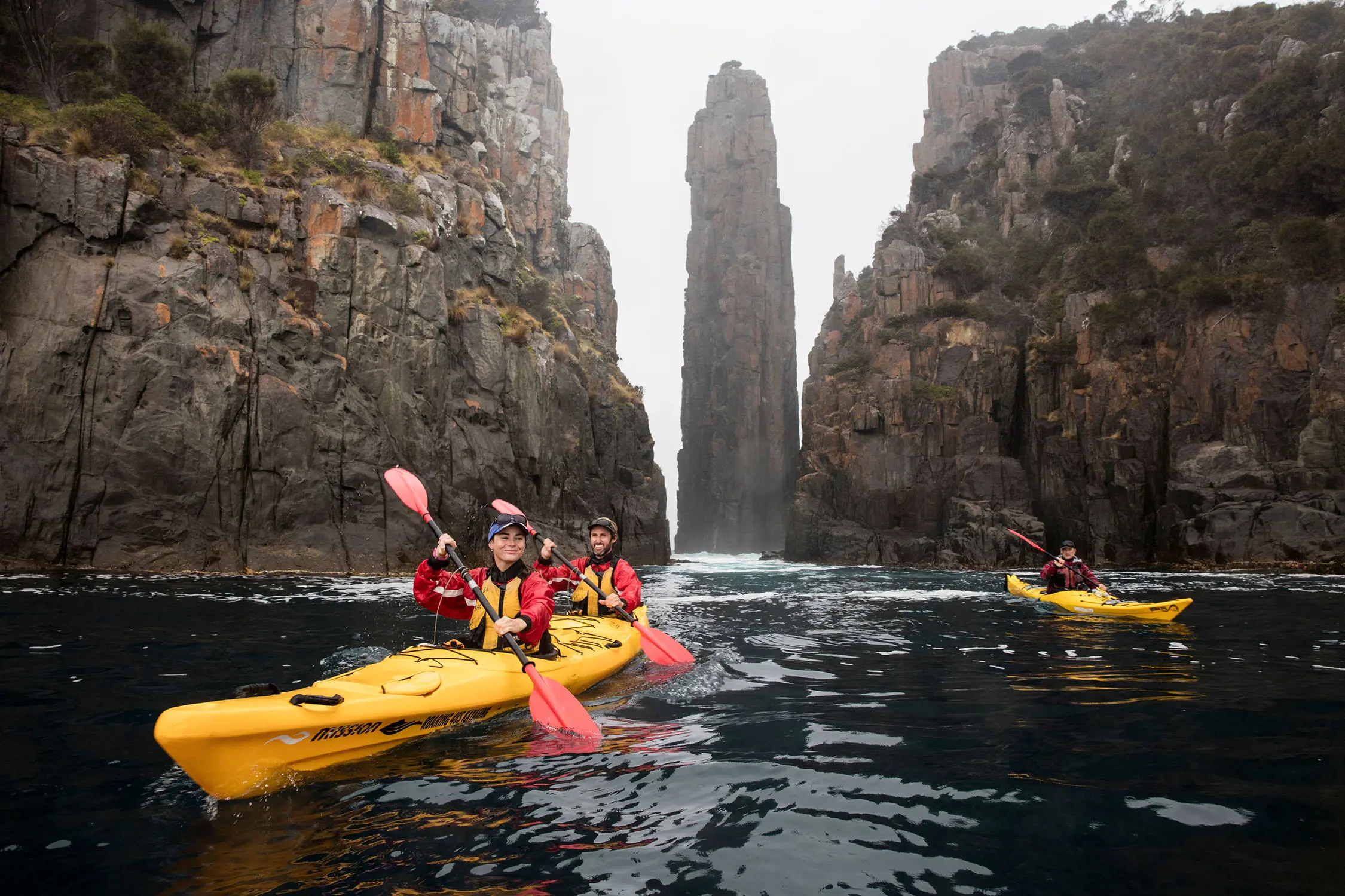 People in yellow kayaks row through dark, still water. They are dwarfed by enormous cliffs either side, and a tall rock pillar behind them.