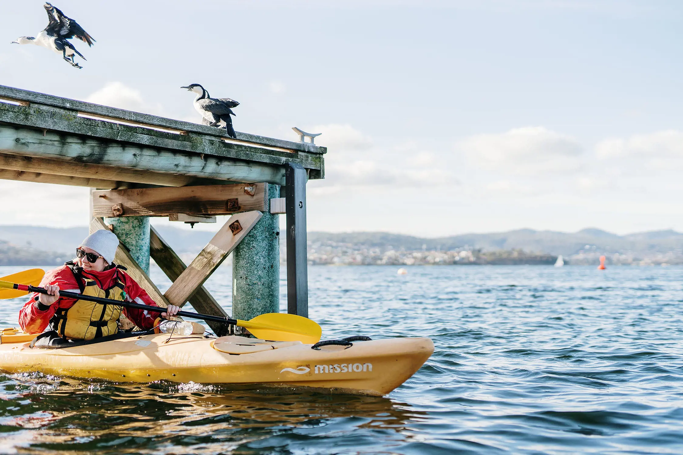 A woman in a yellow kayak paddles underneath an aged jetty. Two birds are mid-flight, landing on the wooden structure above her.