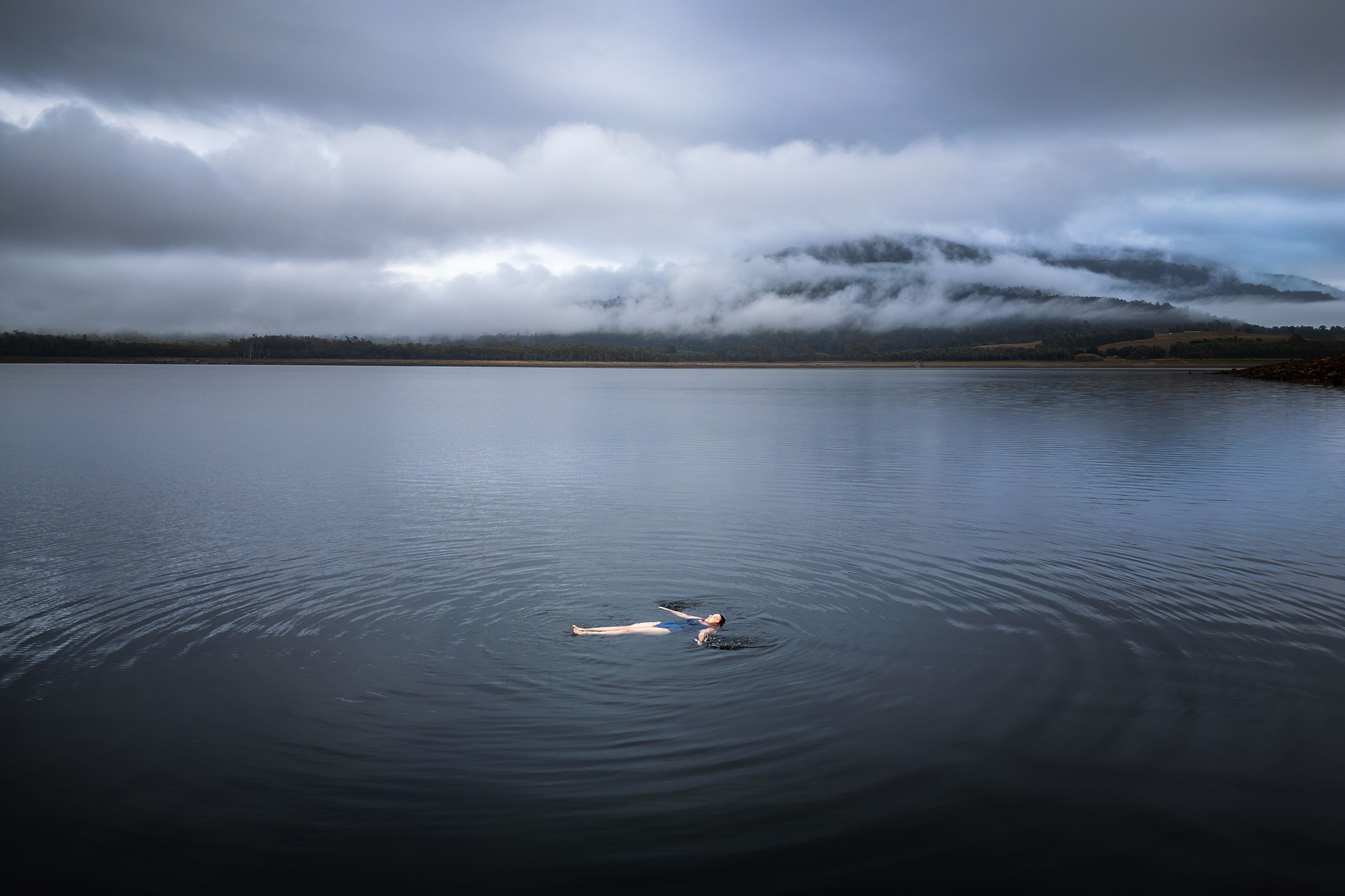 A person floats on their back in a huge, dark lake, with ripples emanating out across the water. Heavy clouds hang low overhead and over mountains in the distance.