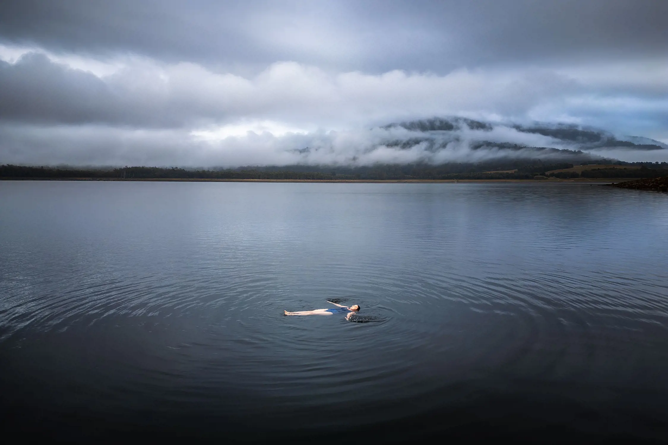 A person floats on their back in a huge, dark lake, with ripples emanating out across the water. Heavy clouds hang low overhead and over mountains in the distance.