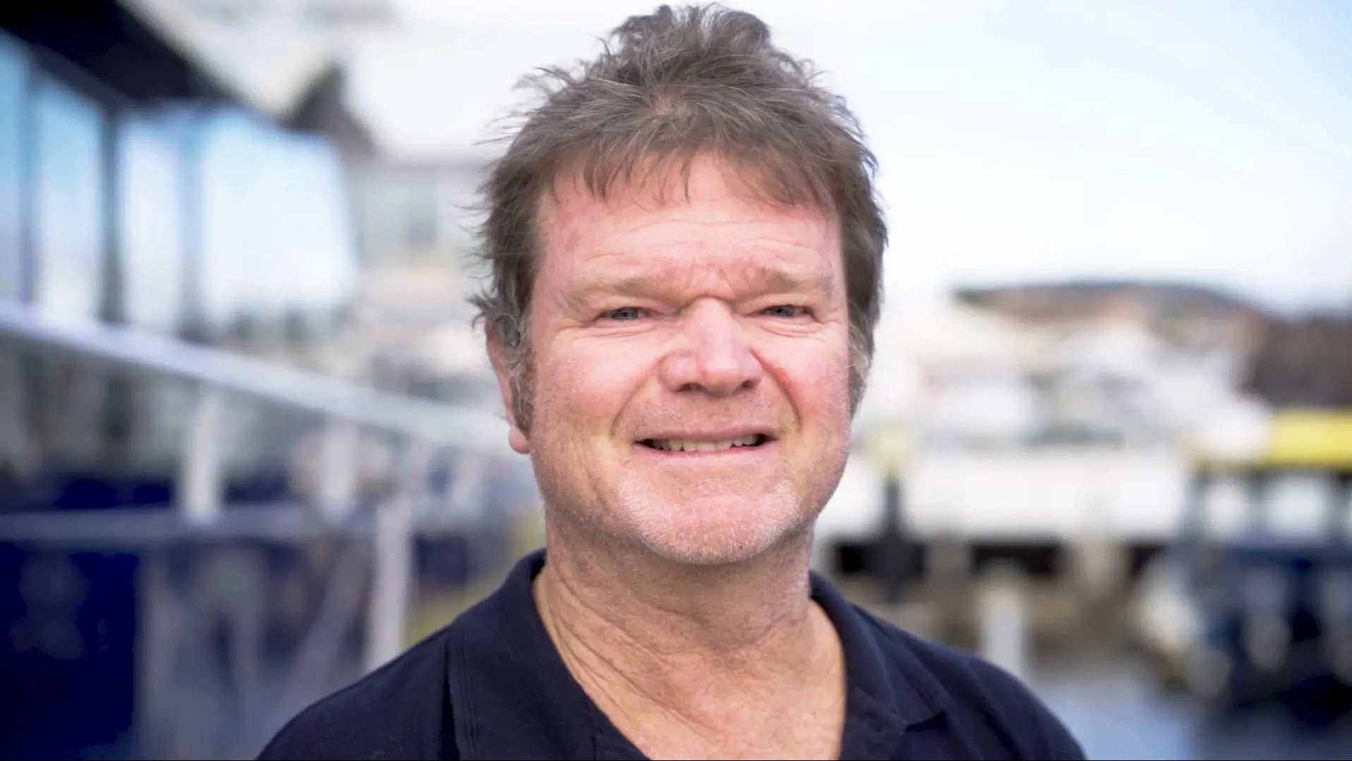 A portrait photo of a man in a navy blue polo shirt, smiling into the camera. Behind him is an out-of-focus wharf scene.